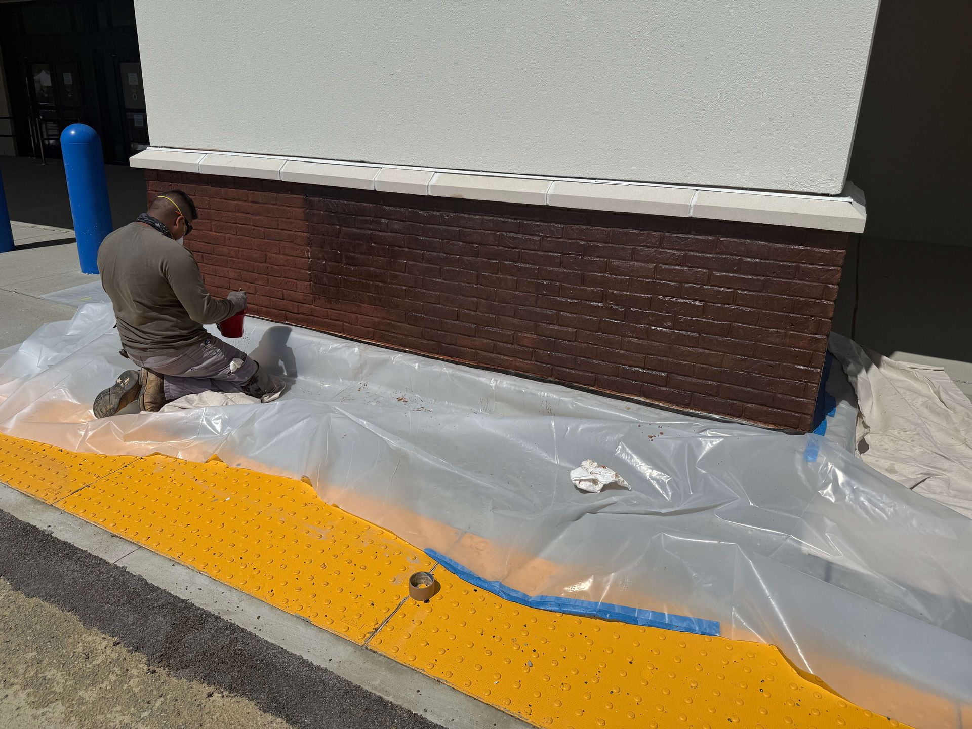 A man is kneeling on the sidewalk painting a brick wall.