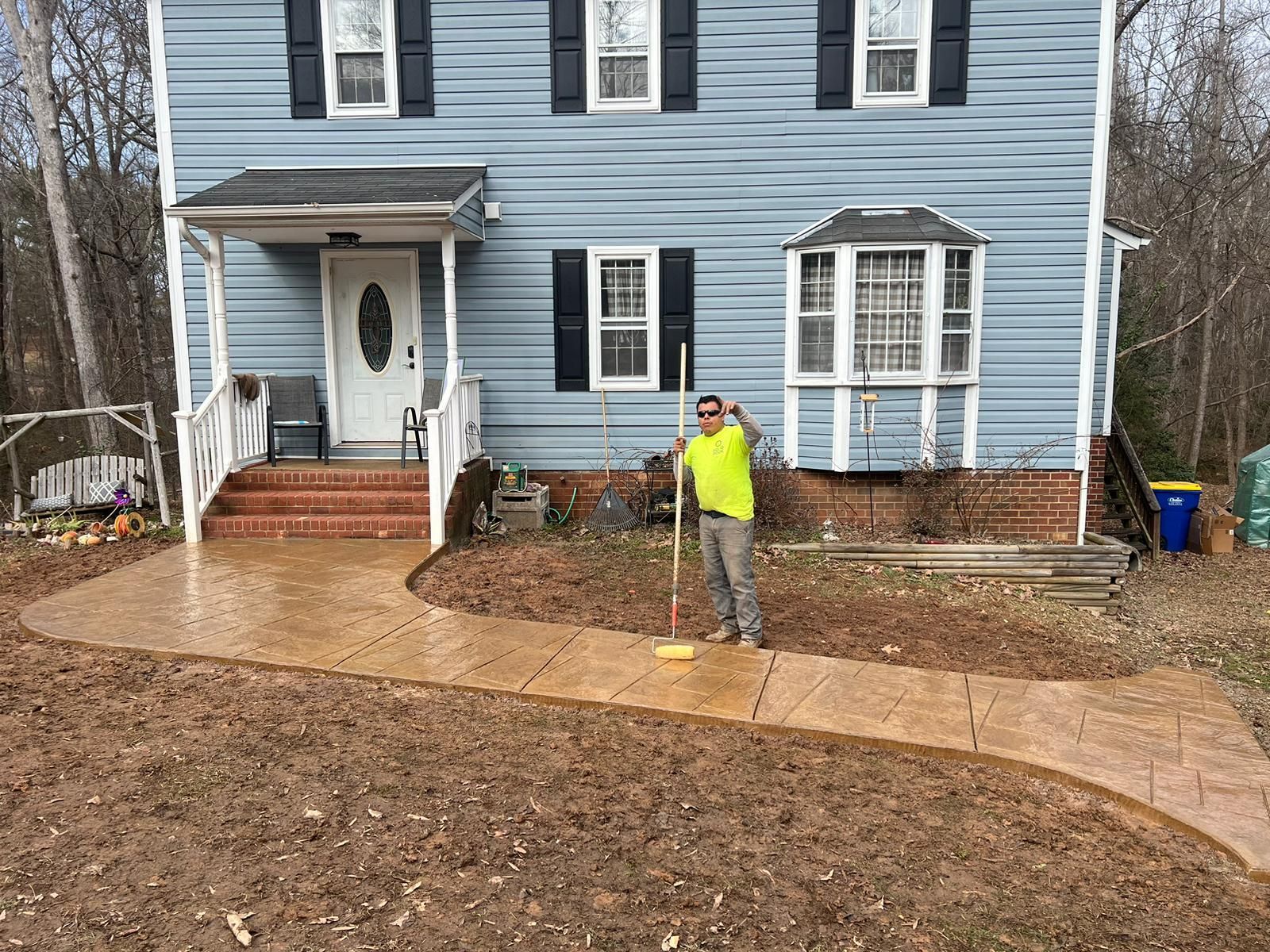 A man is standing on a sidewalk in front of a house holding a broom.