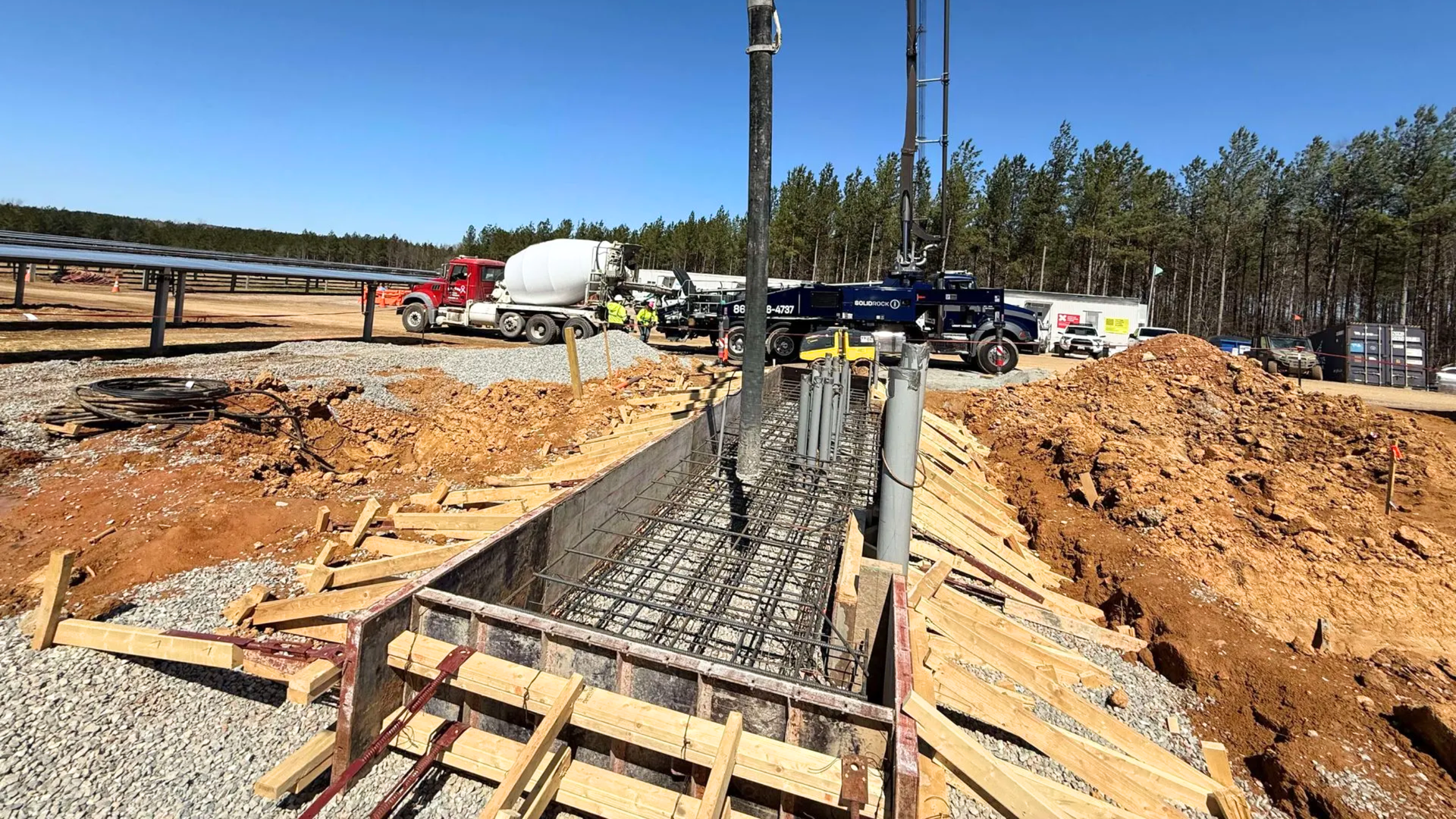 Concrete being poured into a trench by a boom truck at a construction site with several vehicles and solar panels.