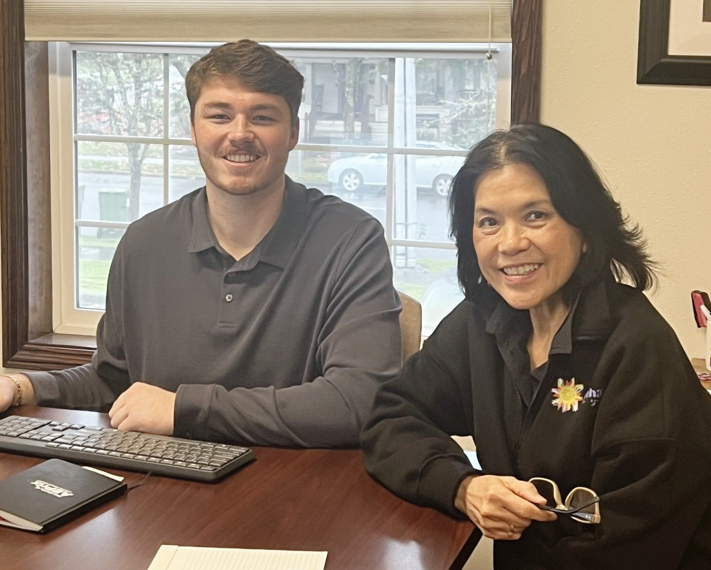 Preston Gullum & Om Sukheenai with Chehalem Insurance Associates in Newberg, Oregon, are sitting at a table and smiling. They are ready to review your insurance policy needs for 2026. 