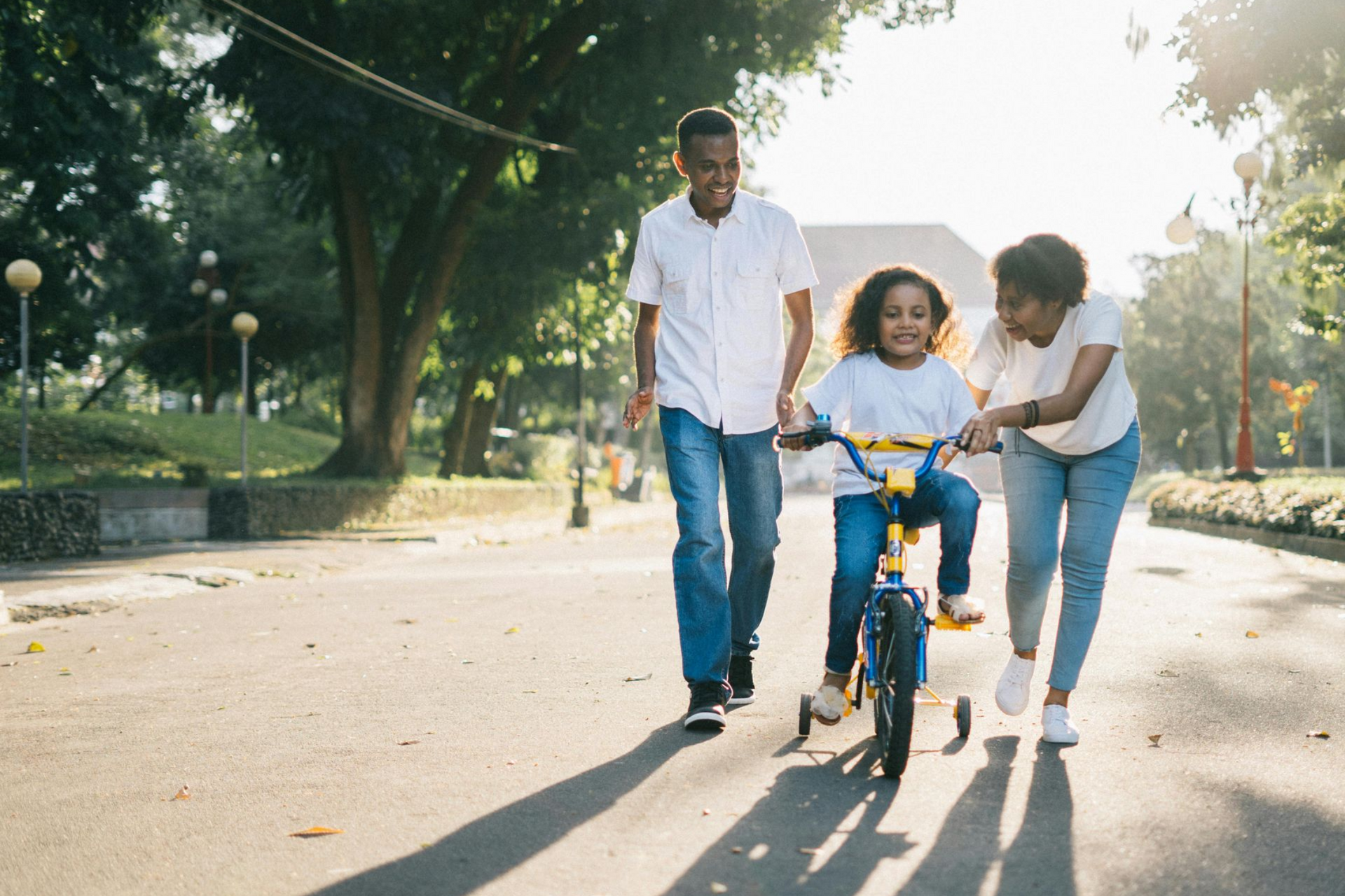Parents teaching a child to ride a bike with training wheels in a park on a sunny day.