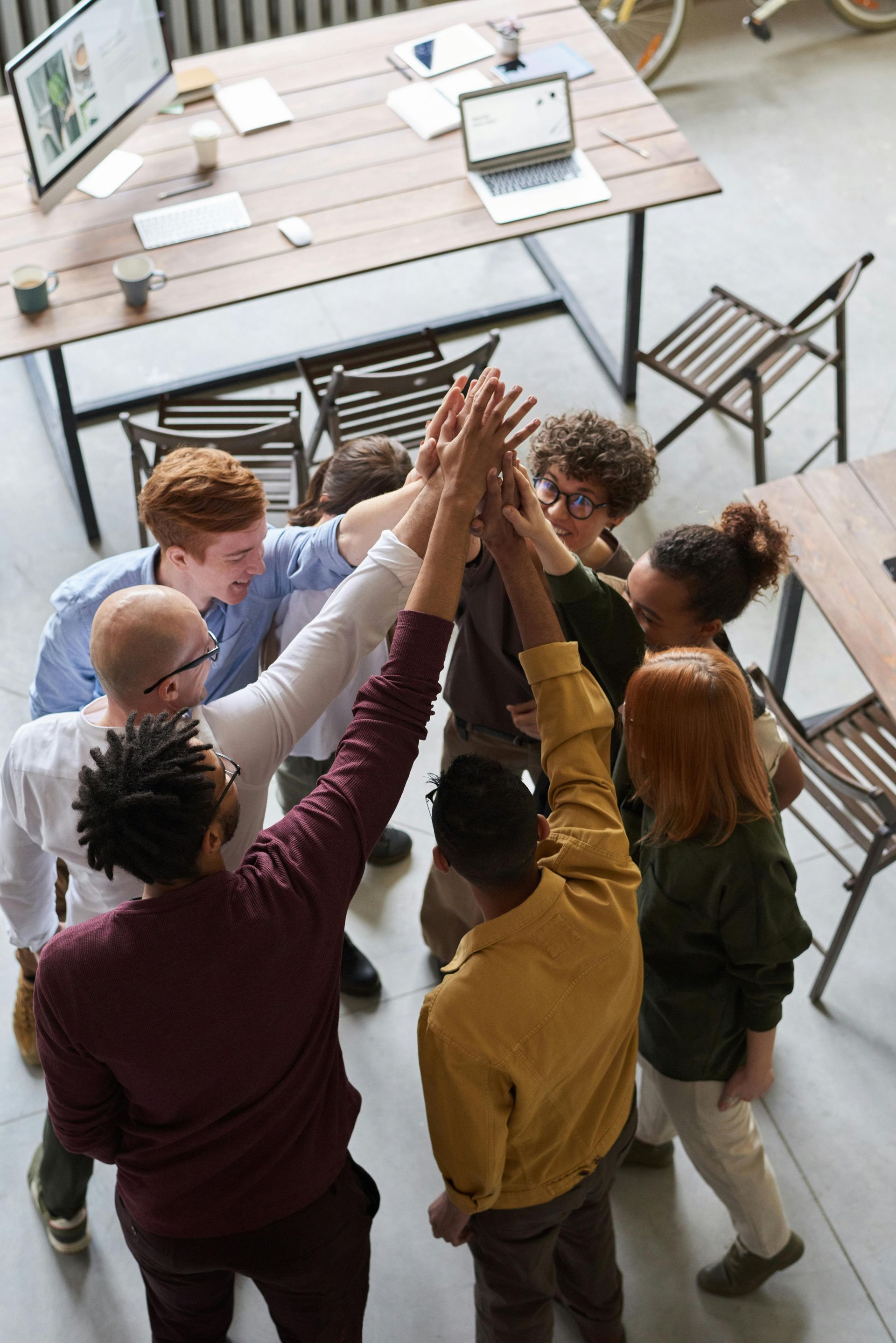 Group of people in a circle, hands raised in a high-five gesture. Wooden table and office setting.