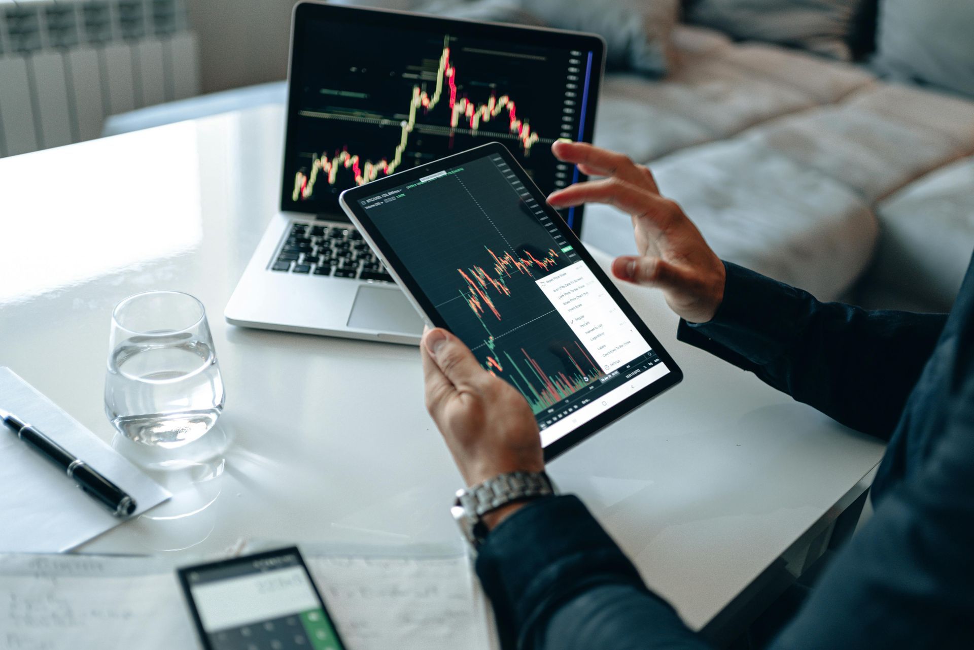 Person analyzing stock charts on a laptop and tablet, white desk, glass of water.