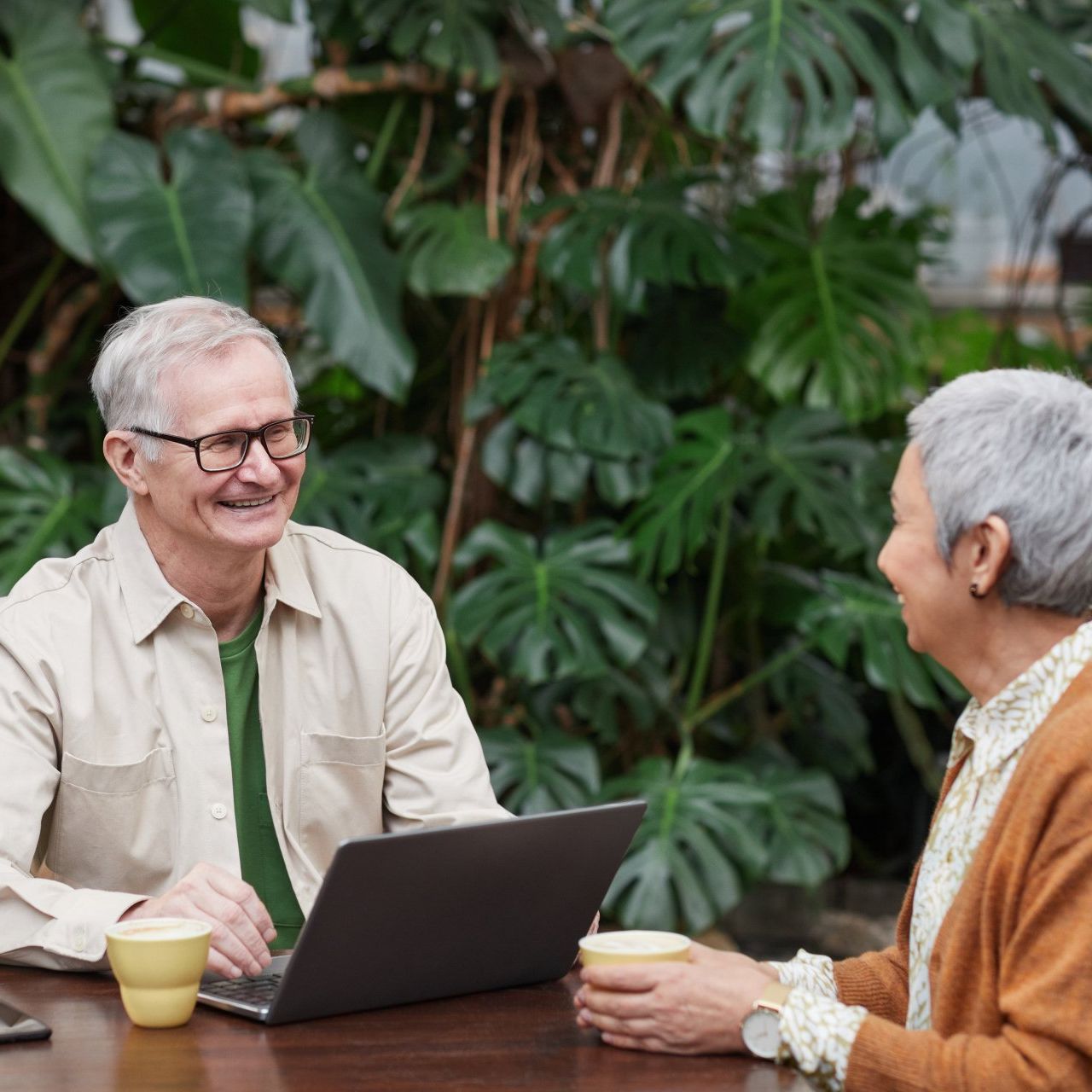 Man and woman seated at table, looking at laptop and smiling, with foliage in background.