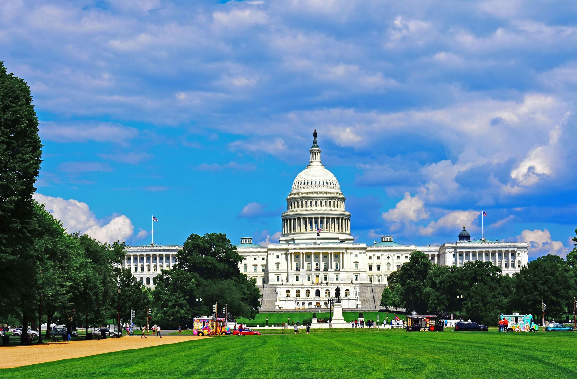 The U.S. Capitol building stands against a bright blue, cloudy sky, overlooking a green lawn and park area.