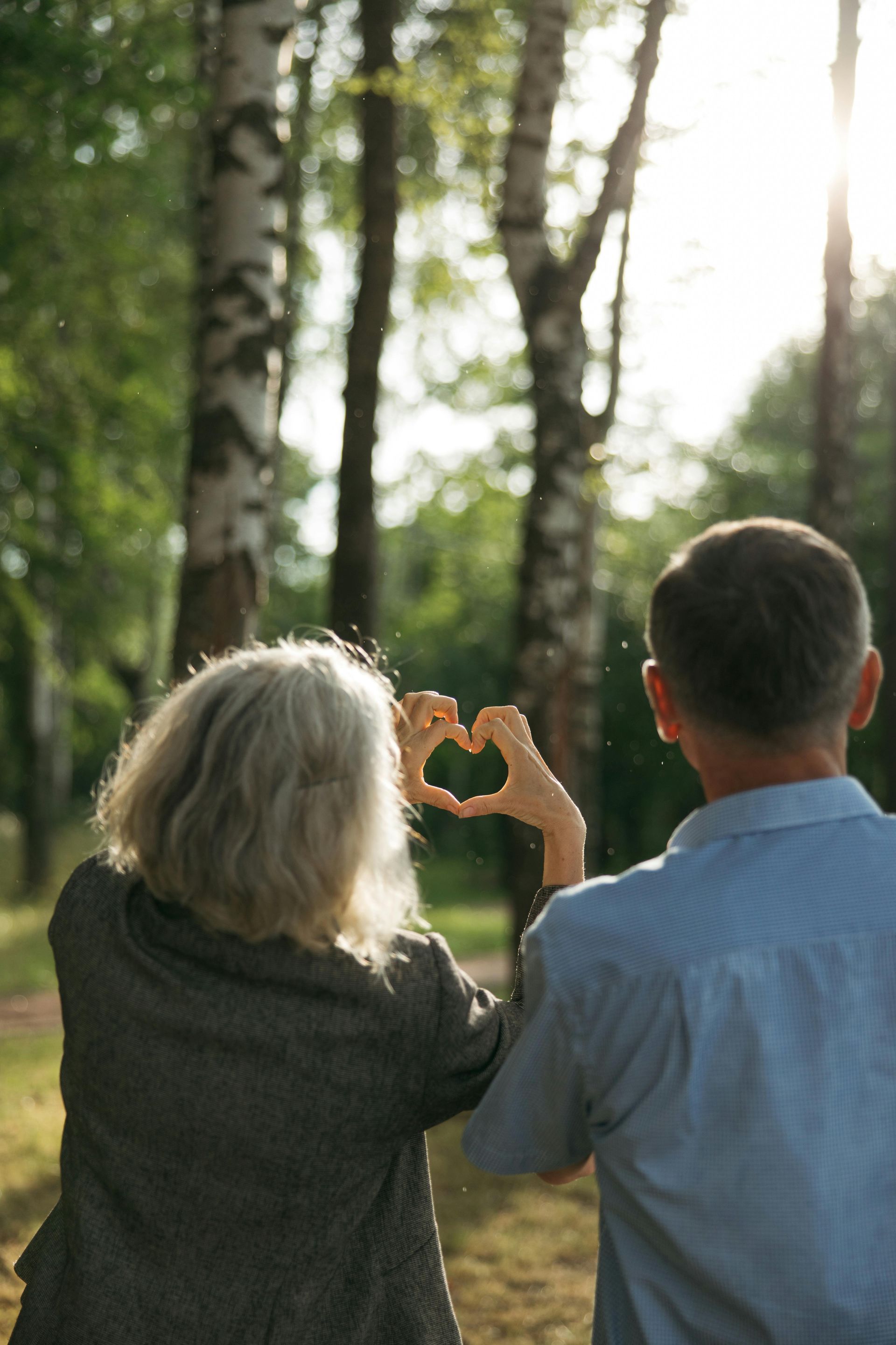 Couple outdoors, forming a heart with their hands against a sunlit forest background.