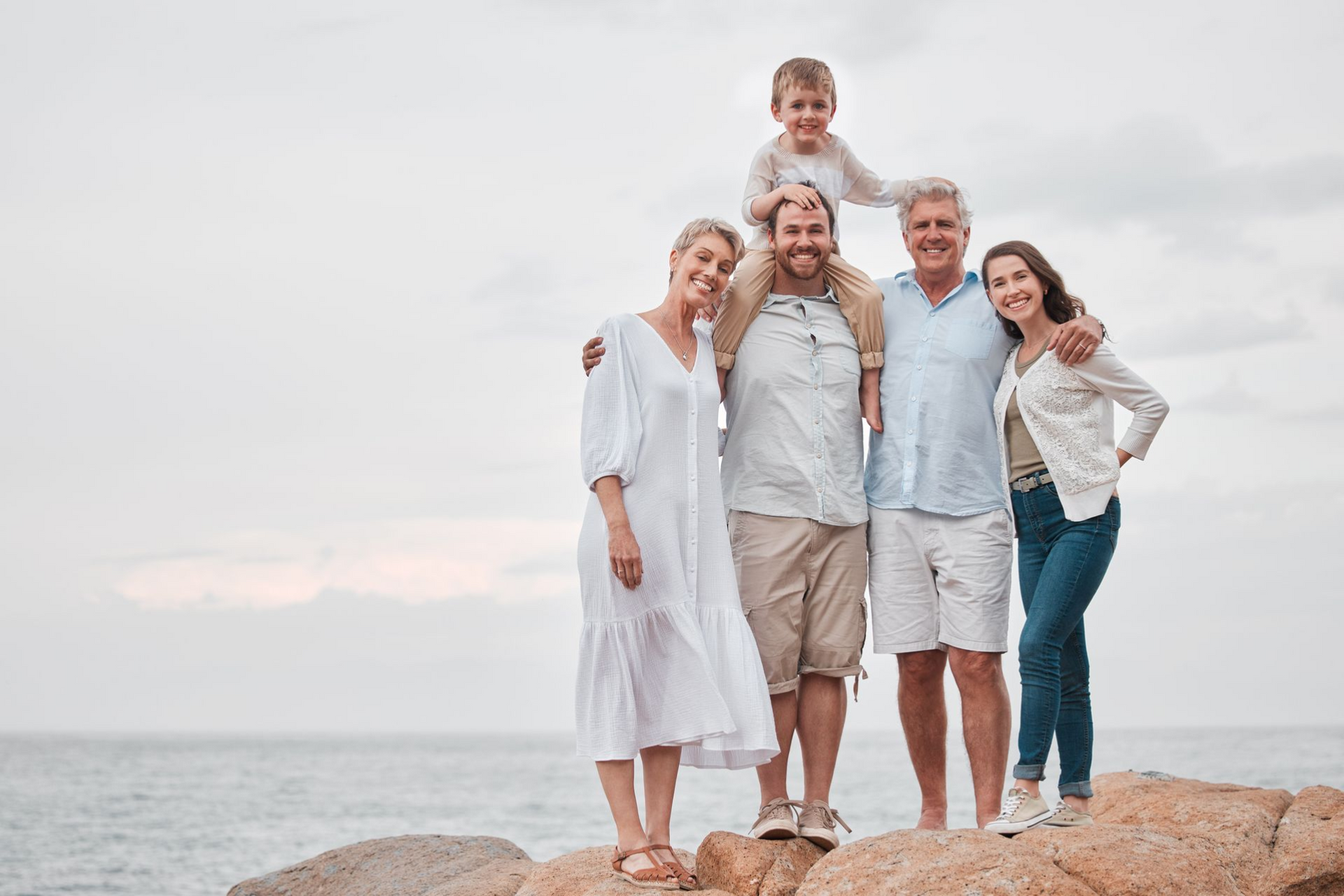 Family of five on rocks by the ocean, smiling. Child on shoulders. Cloudy sky.