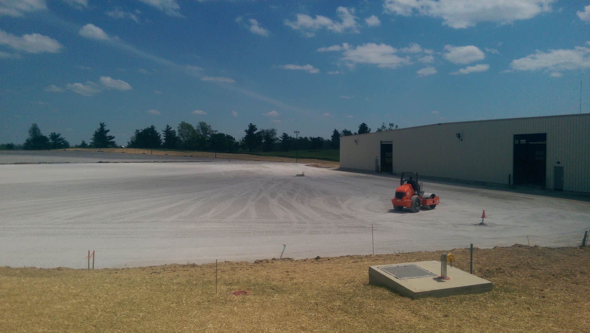 A roller compacting a light-colored surface, possibly concrete, next to a white industrial building.