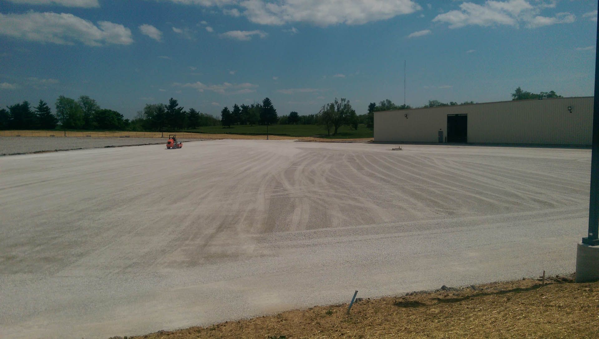 A large, light-colored gravel lot with a building on the right, trees in the background, under a blue sky.