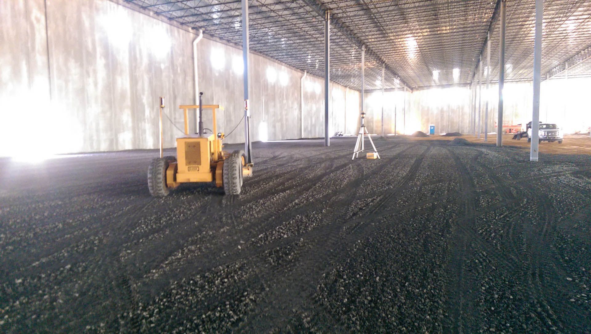 Yellow grader smoothing dark surface inside a large, unfinished building.