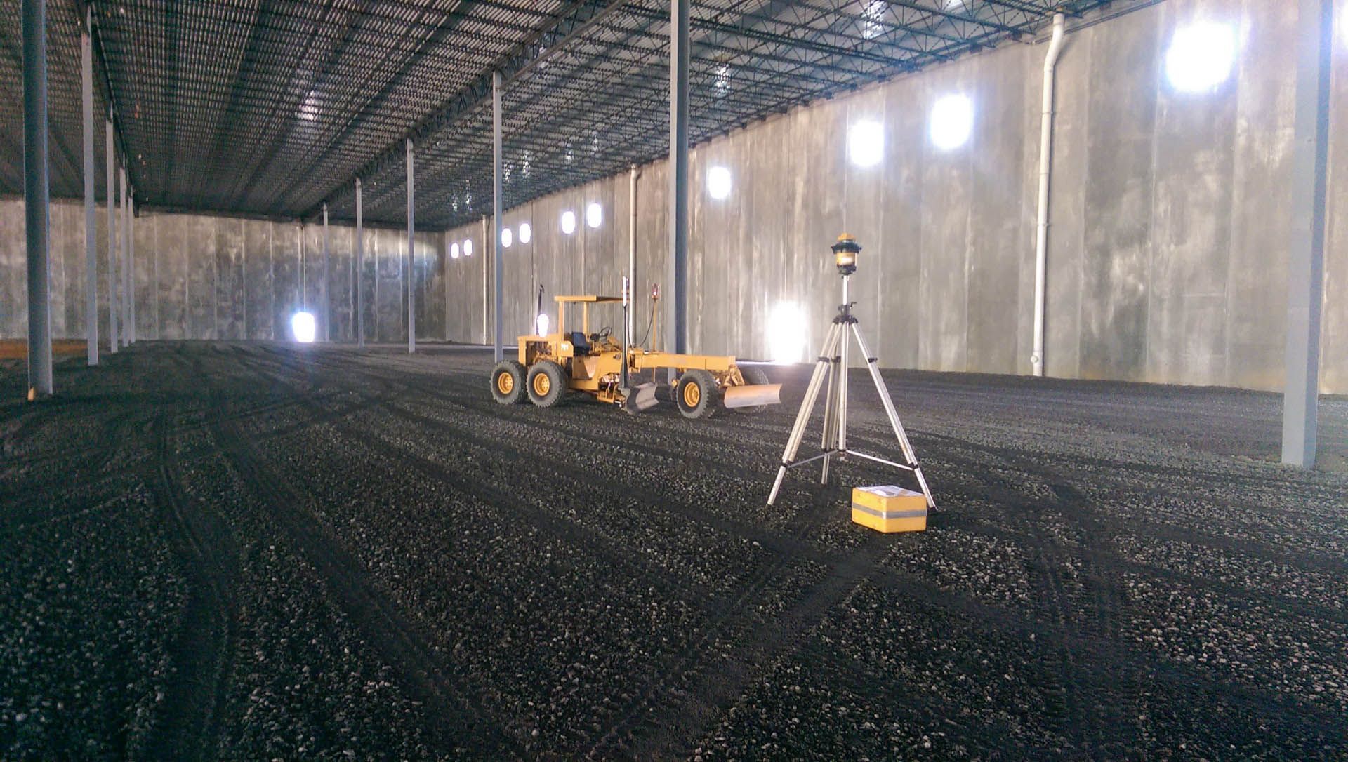 Construction site interior: a yellow grader, surveying equipment, and a level dark ground, under a steel-framed ceiling.