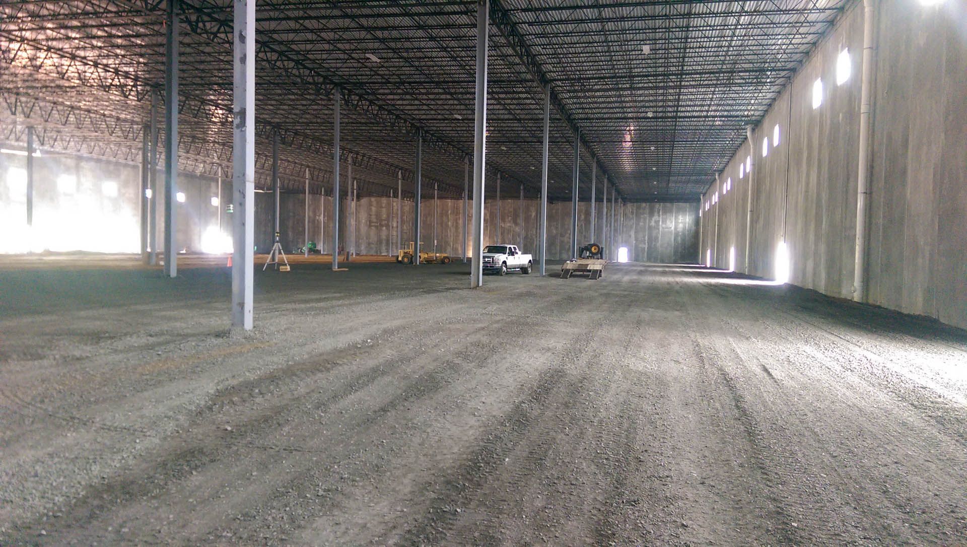 Inside a large warehouse; gravel floor, metal beams, cement walls, daylight through openings, vehicles.
