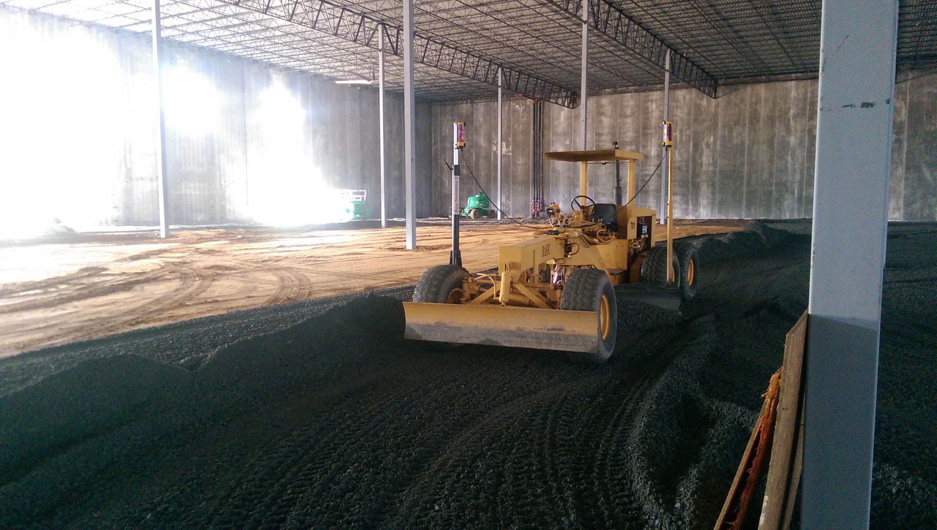 A yellow grader leveling dark gravel inside a large, open metal building.