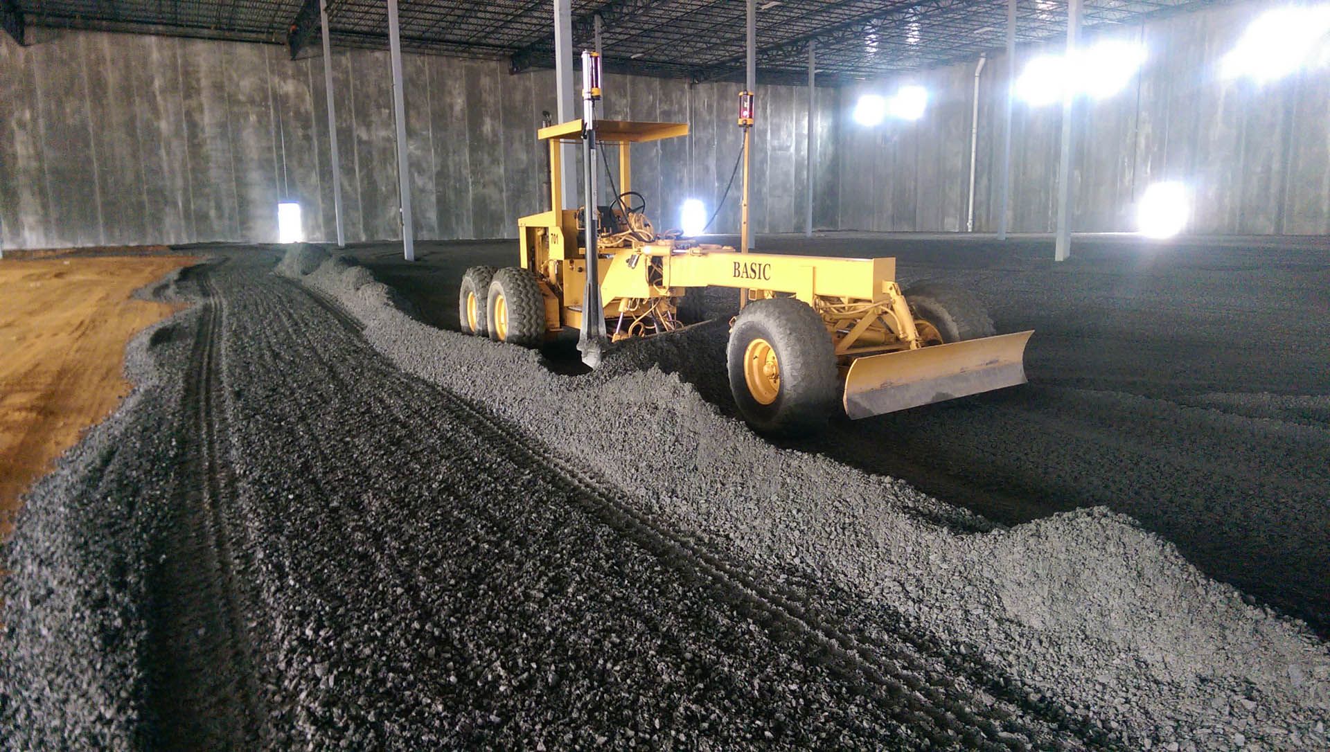 A yellow motor grader smoothing dark gravel inside a large indoor arena.