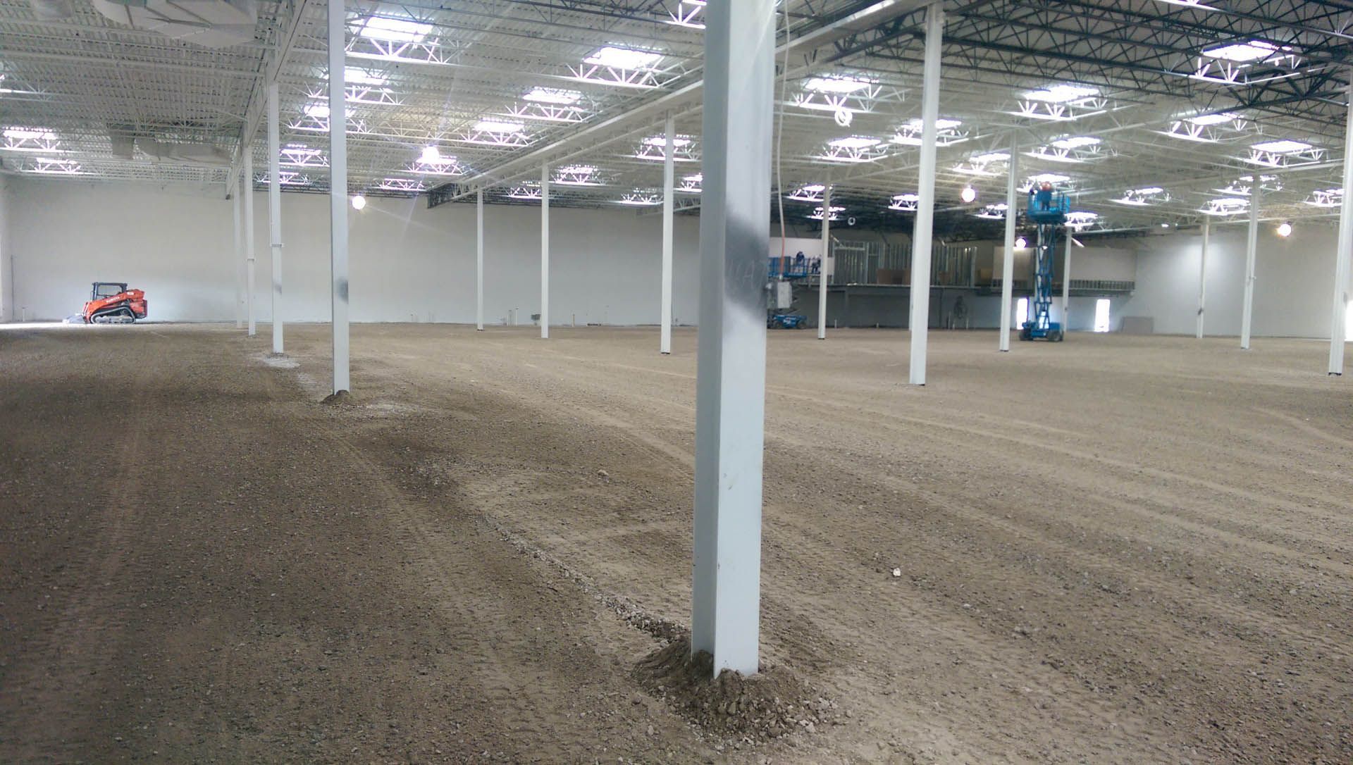 Large empty warehouse with dirt floor, white support columns, and construction equipment.