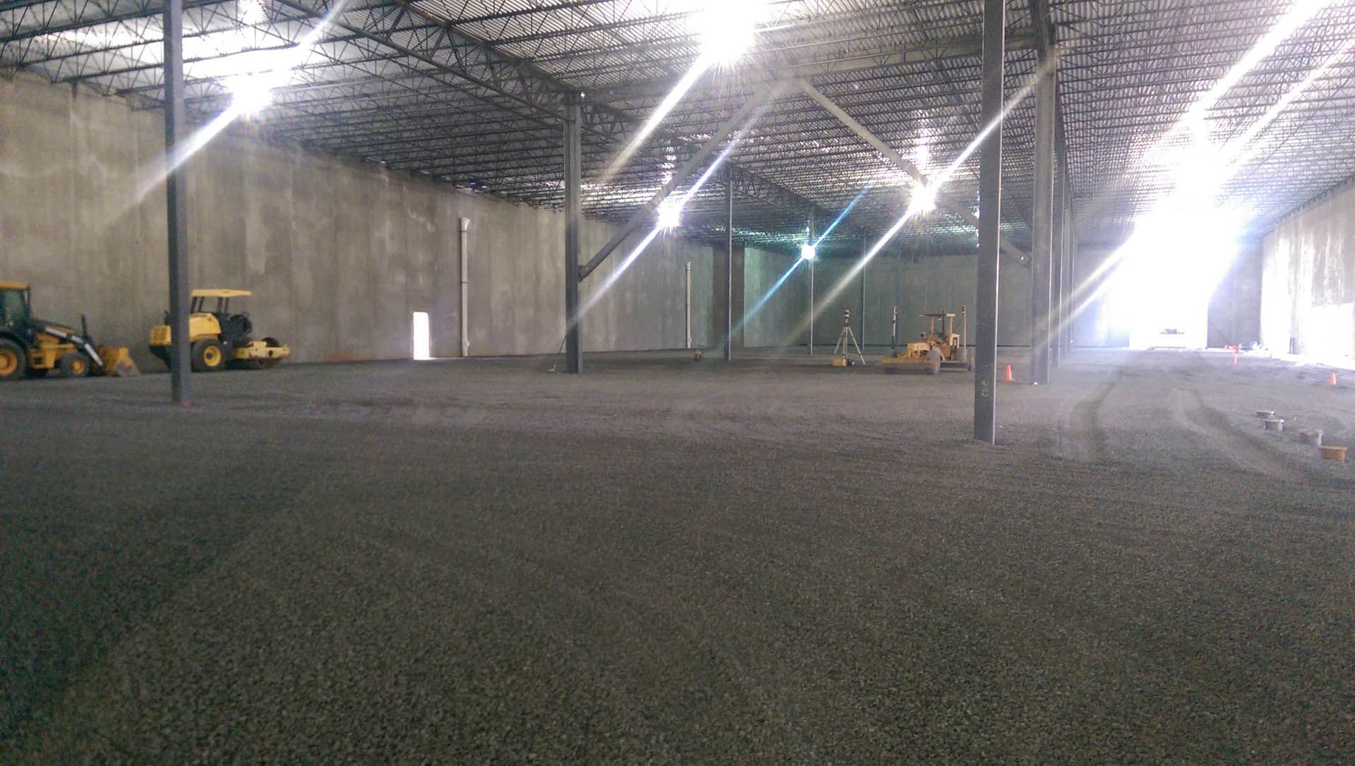 Interior of a large warehouse with a gravel floor, construction equipment, and bright overhead lights.