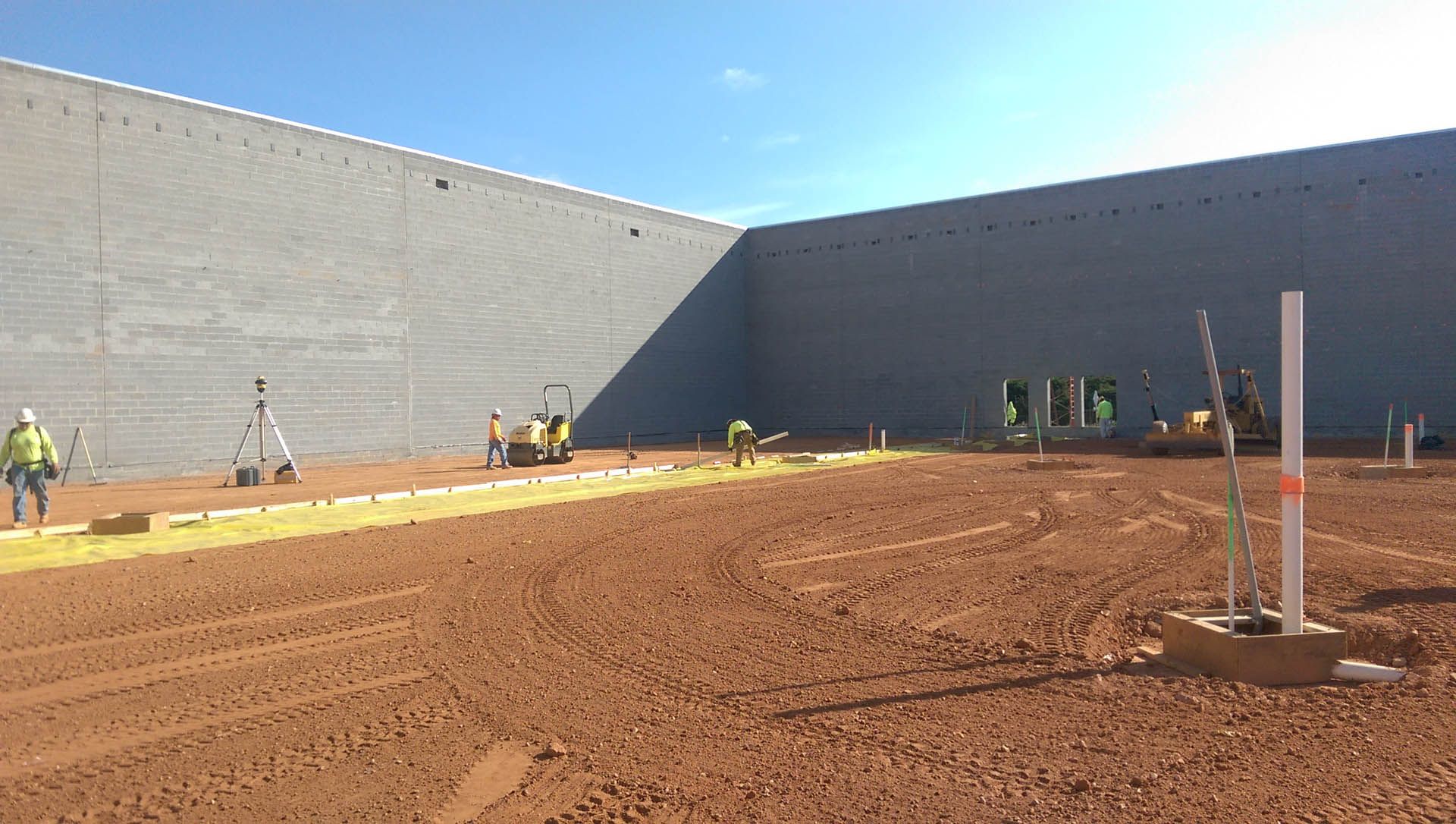 Construction site with workers, dirt, and a building under construction. Sunny, blue sky.