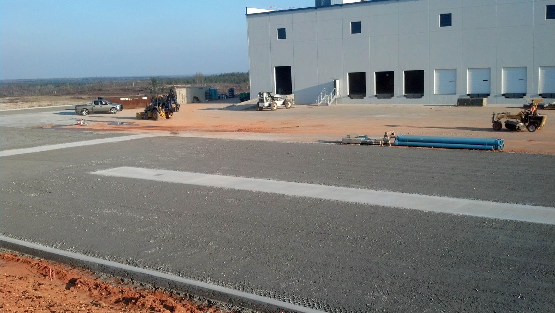 Construction site with a gray concrete road, heavy machinery, and a large white industrial building.