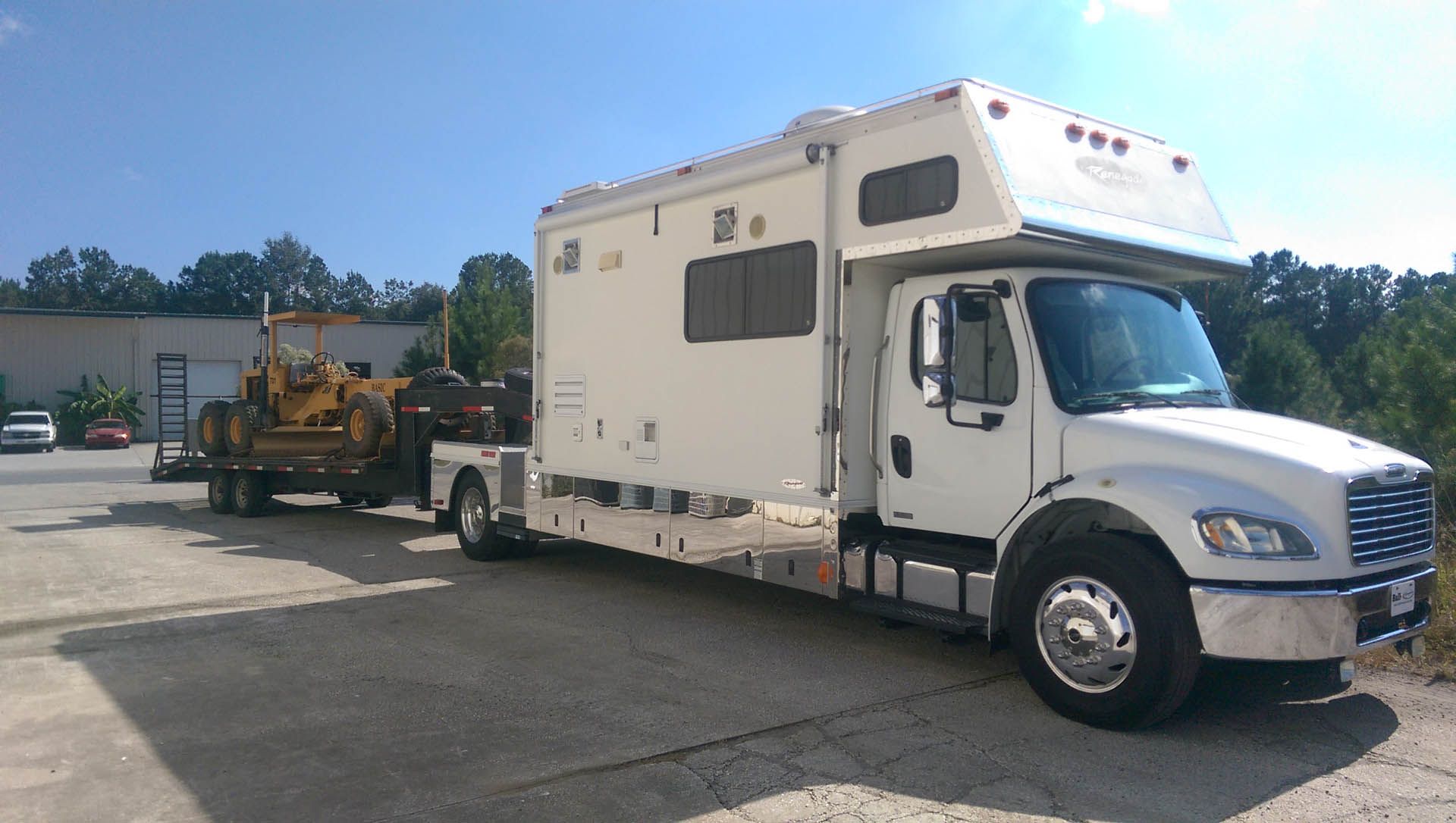 White RV truck towing a trailer with yellow bulldozer parked outside on a sunny day.