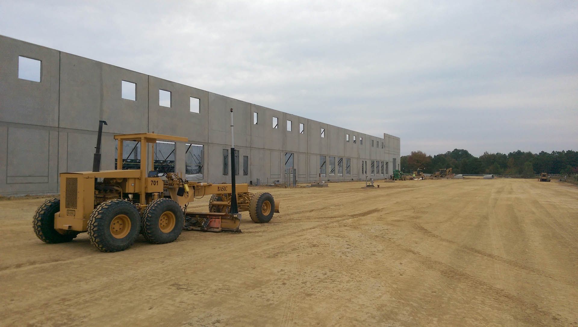 A yellow grader levels dirt in front of a concrete building under construction on a cloudy day.
