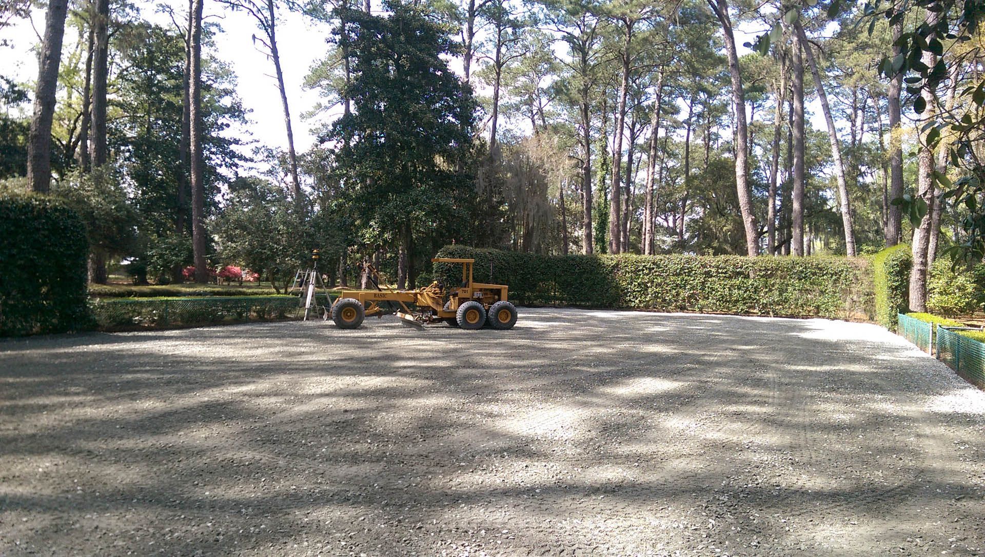 Yellow grader leveling a gravel surface in a park-like setting with trees and hedges.