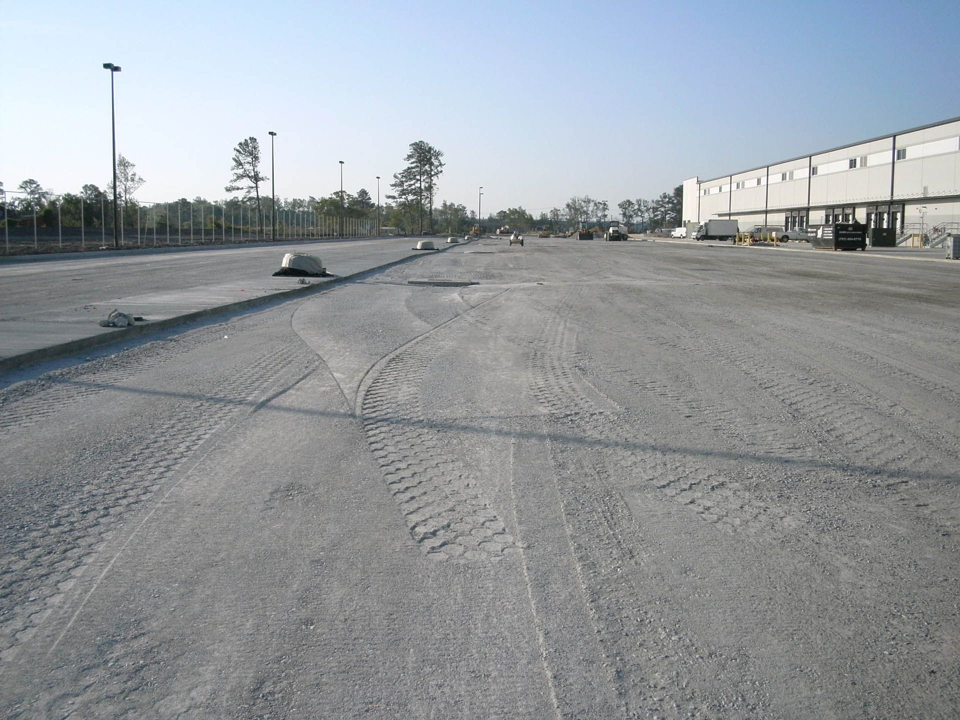 Gravel parking lot near industrial buildings with tire tracks and some trees.