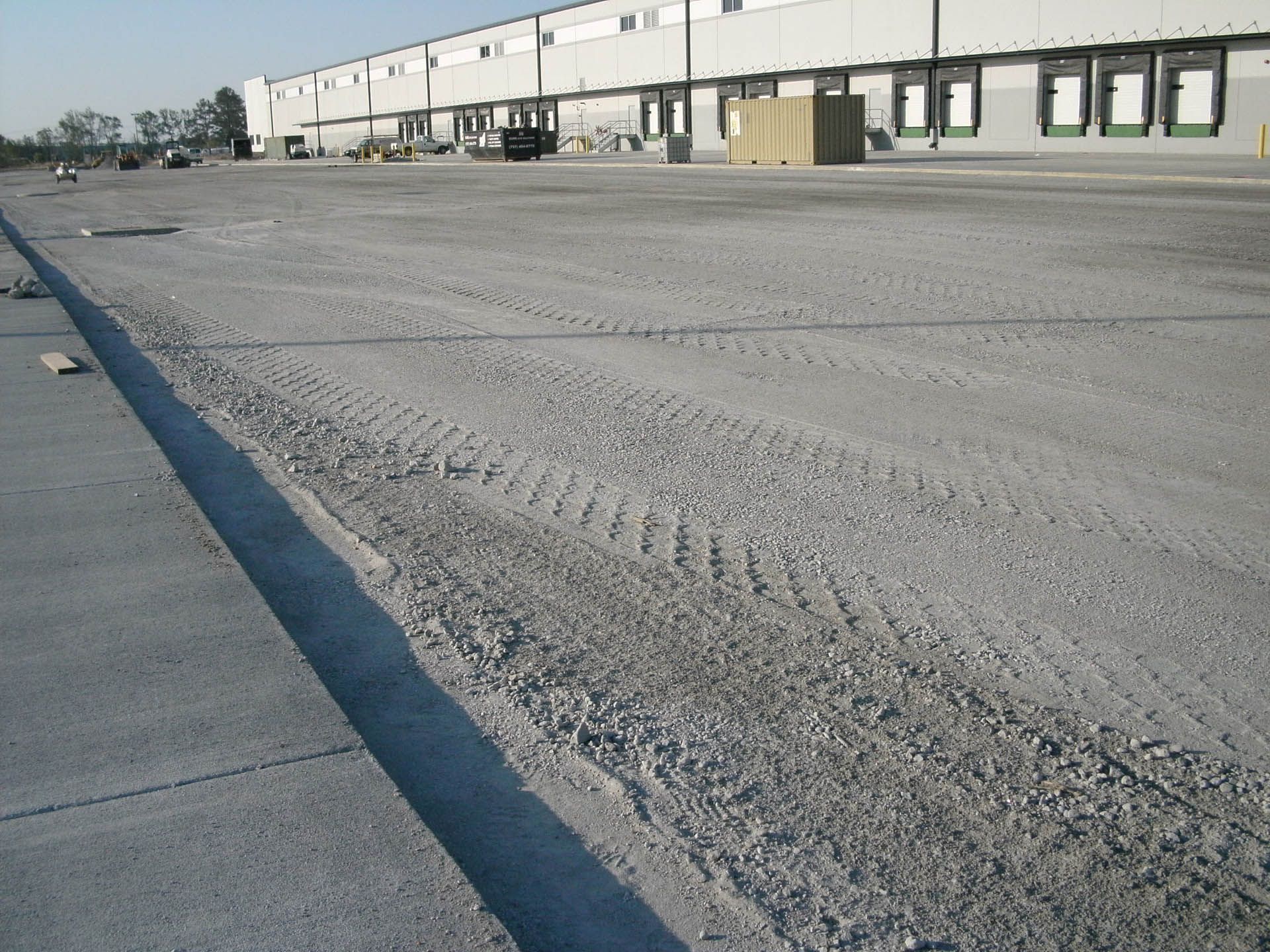 A wide, gravel parking lot with tire tracks, adjacent to a large commercial building with loading docks.
