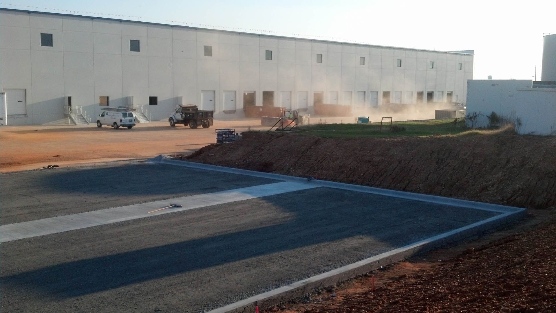 Construction site with a long, white warehouse.  Workers and vehicles in operation.  Gravel and dirt in the foreground.