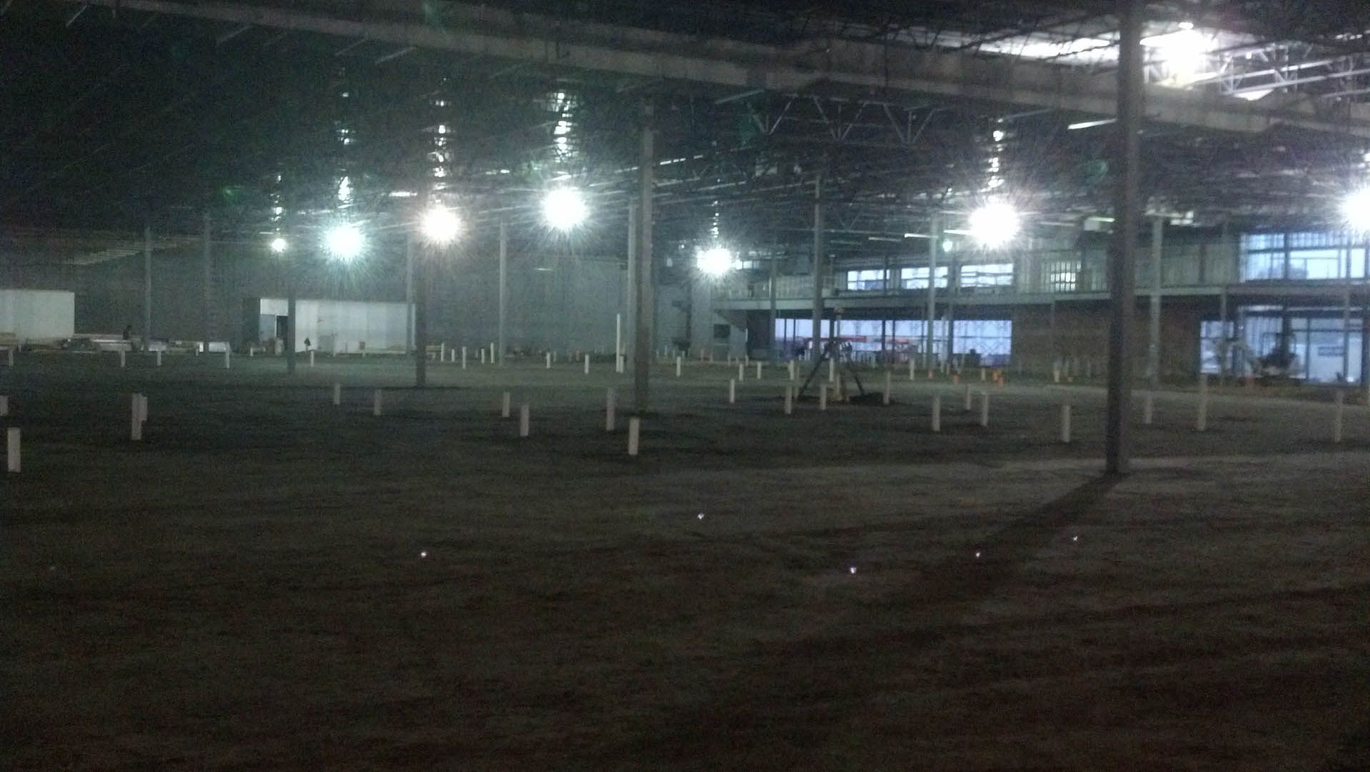 Interior of a large, dimly lit, empty warehouse. Bright overhead lights illuminate the dirt floor and support beams.