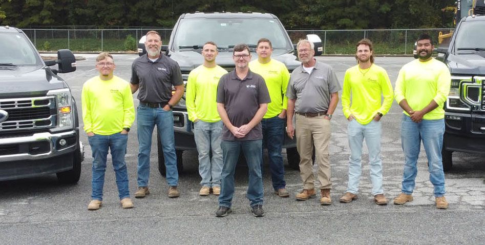 Group of men, some in safety vests, standing by dark trucks, smiling, in a parking lot.
