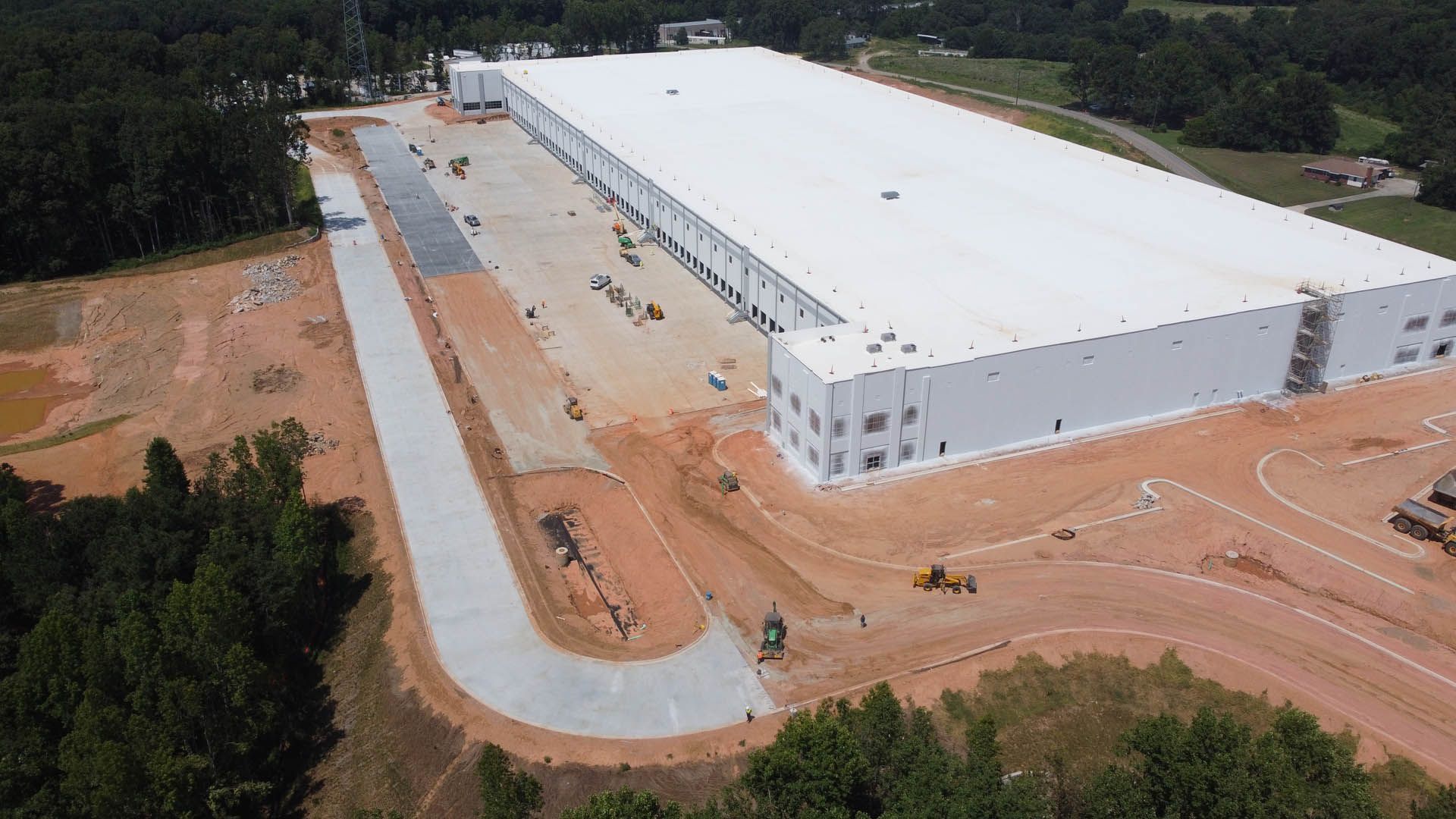 Aerial view of a large, white warehouse under construction. Asphalt road and dirt surroundings.