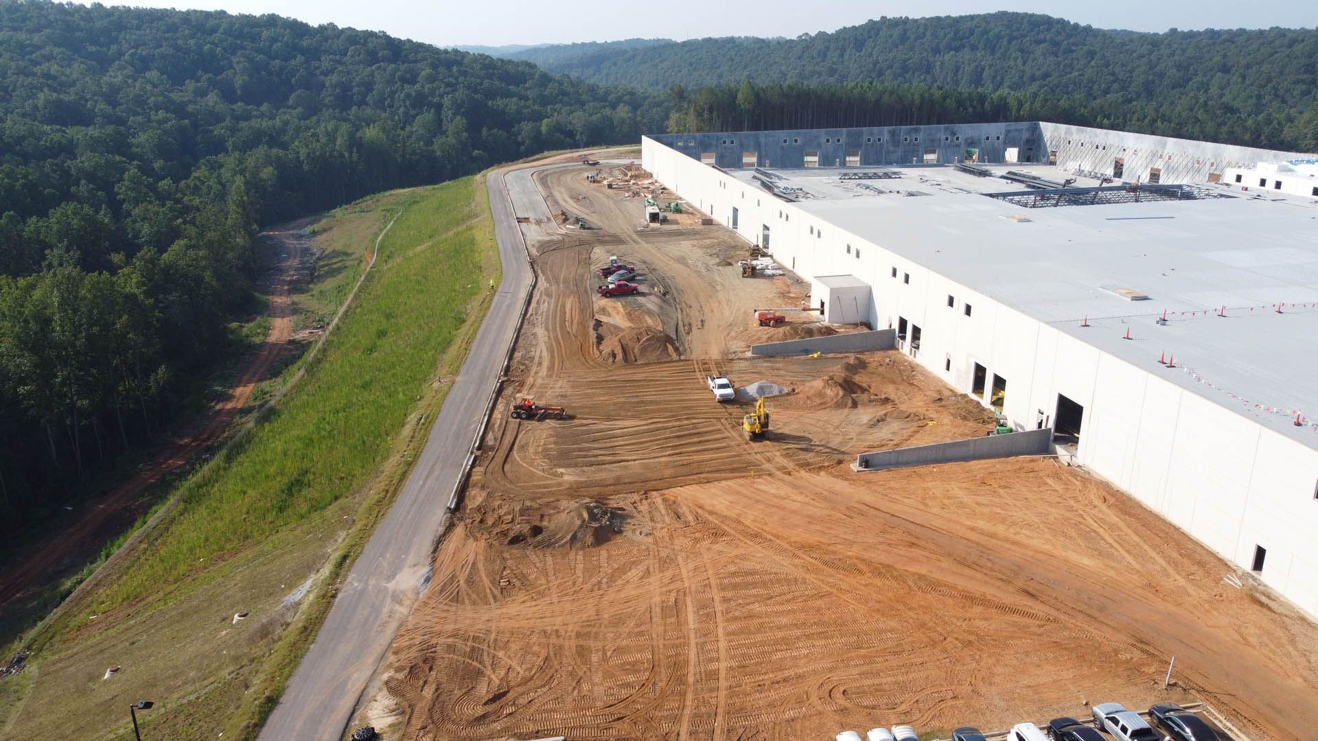 Construction site with a large concrete building, heavy machinery, and a dirt road. Surrounded by trees and hills.