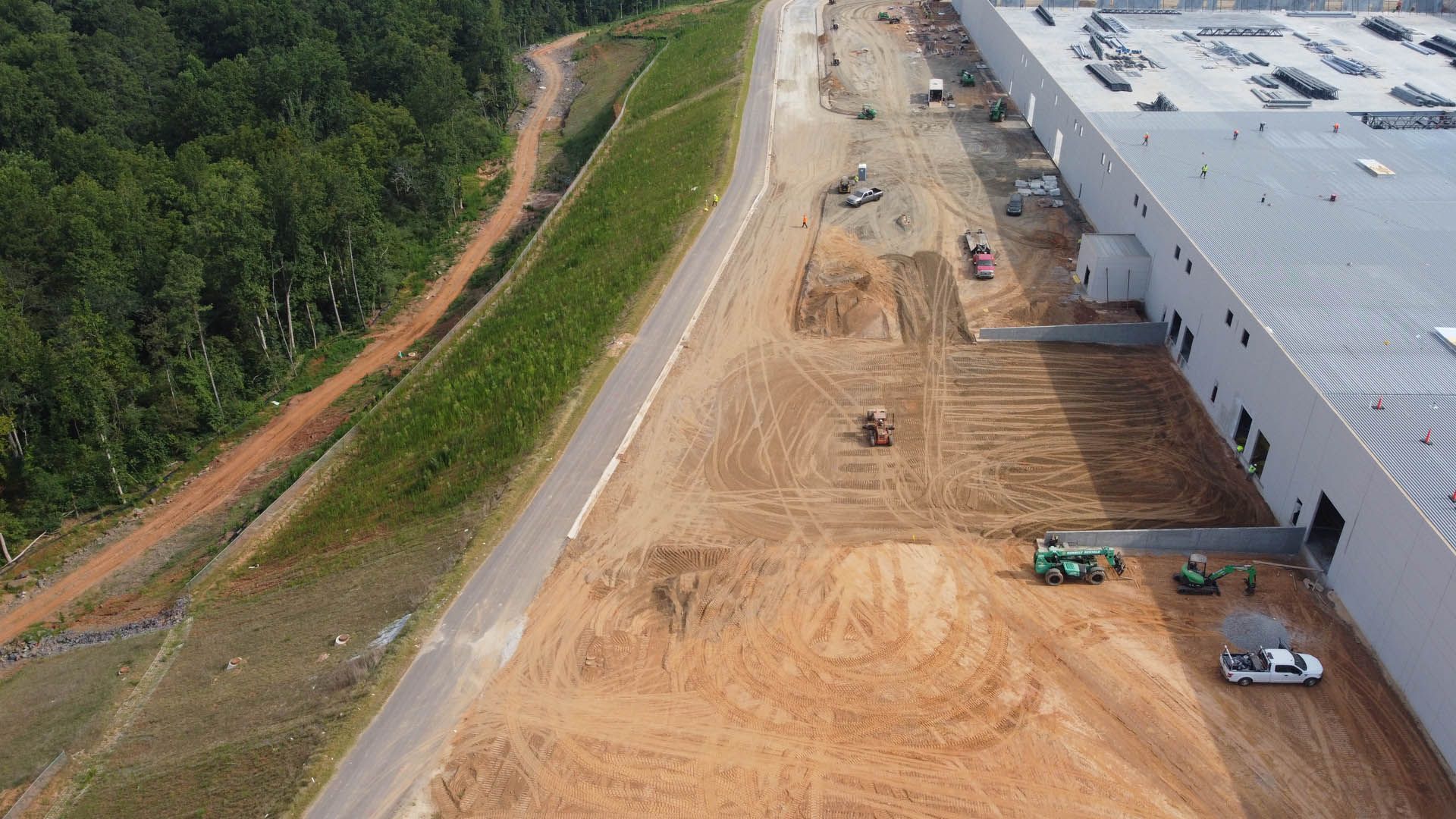 Construction site with a large industrial building. Bulldozers, workers, and grading dirt near a new warehouse.