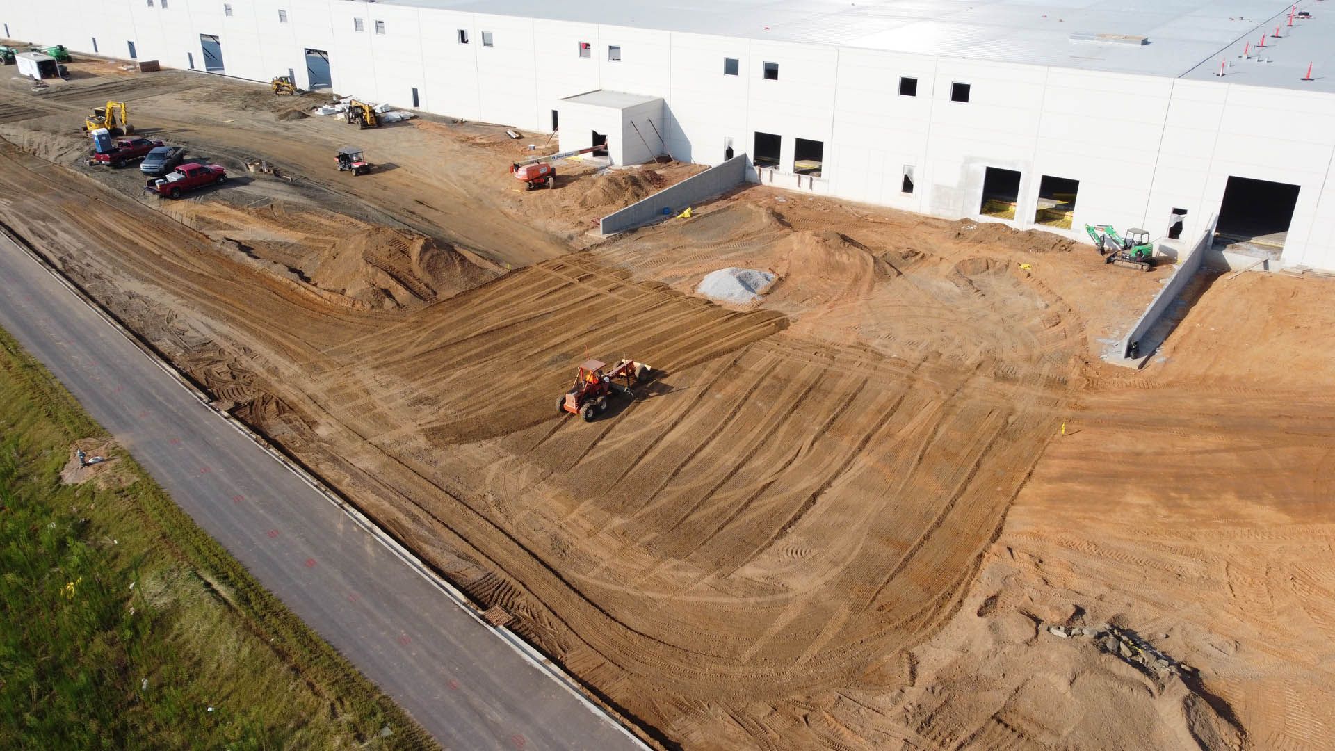 Construction site with heavy machinery grading the brown dirt in front of a white industrial building.