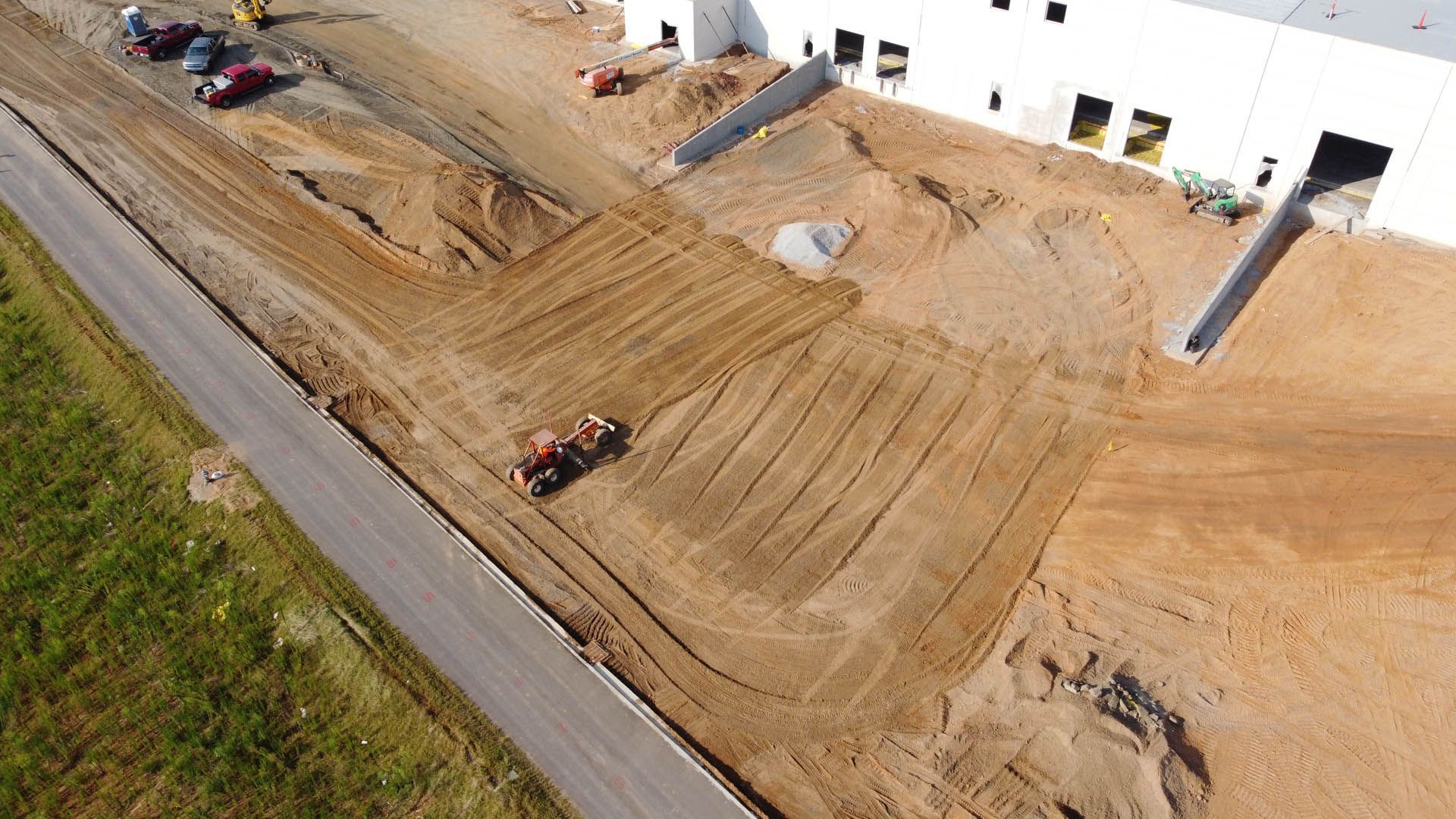 Aerial view of construction site with building, tractor grading soil, and asphalt road.