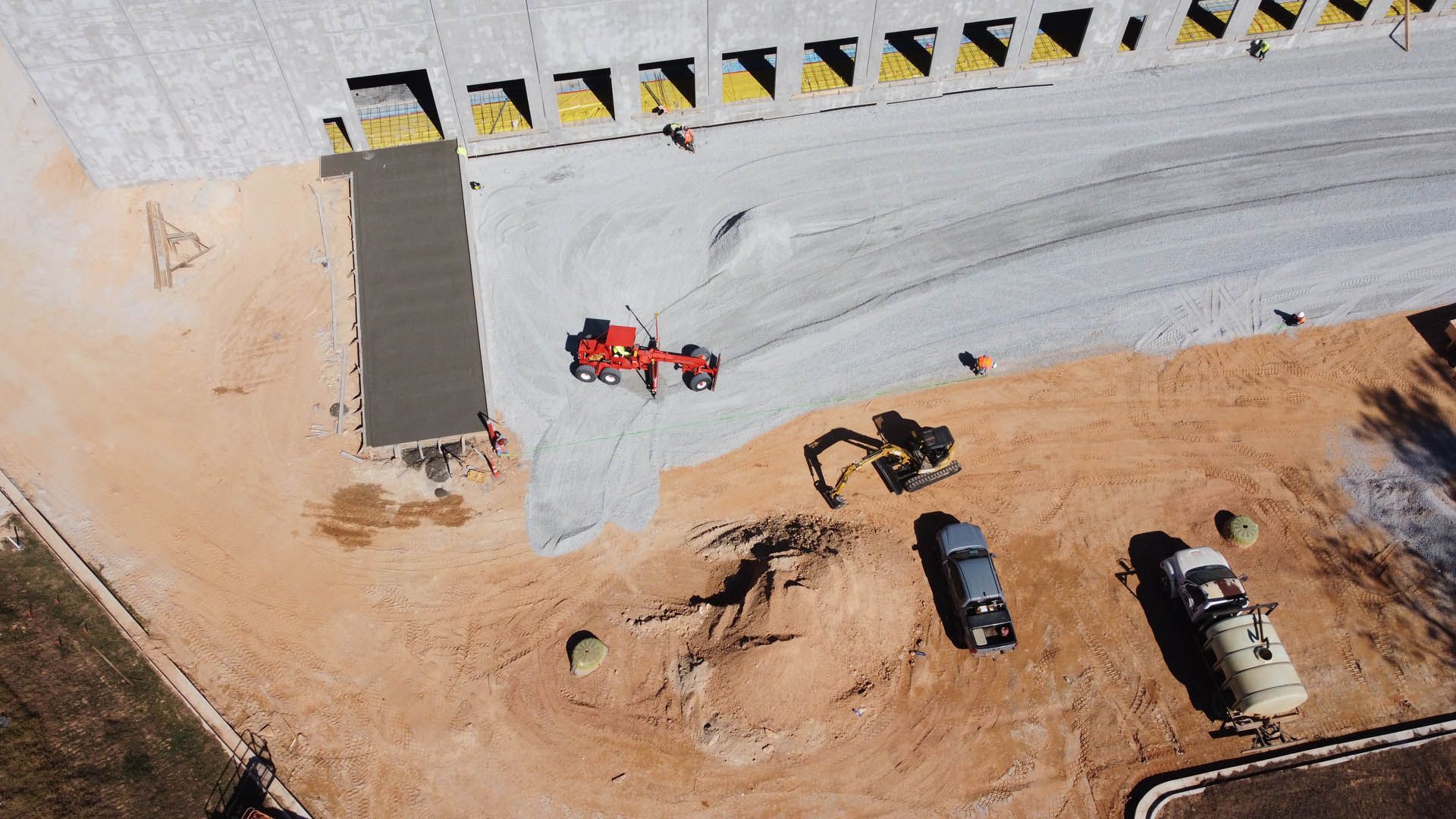 Construction site; tractor smoothing concrete, excavator, trucks, and building in background.