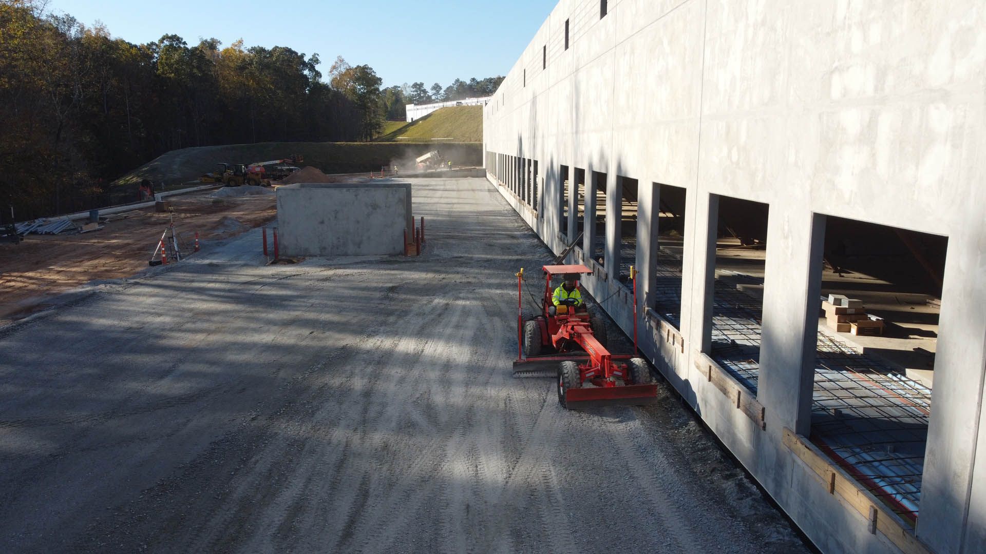 A worker on a red tractor clearing a gravel area near a large concrete building with loading dock doors.