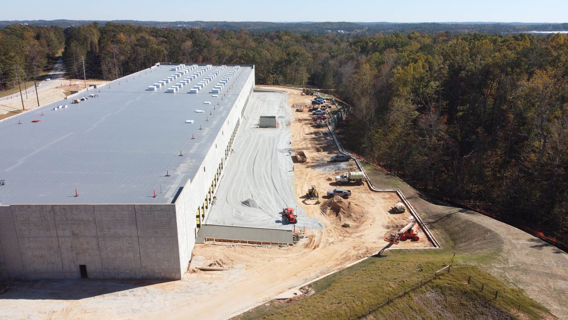 Construction of a long, gray industrial building with a flat roof. Dirt and trees surround the structure.