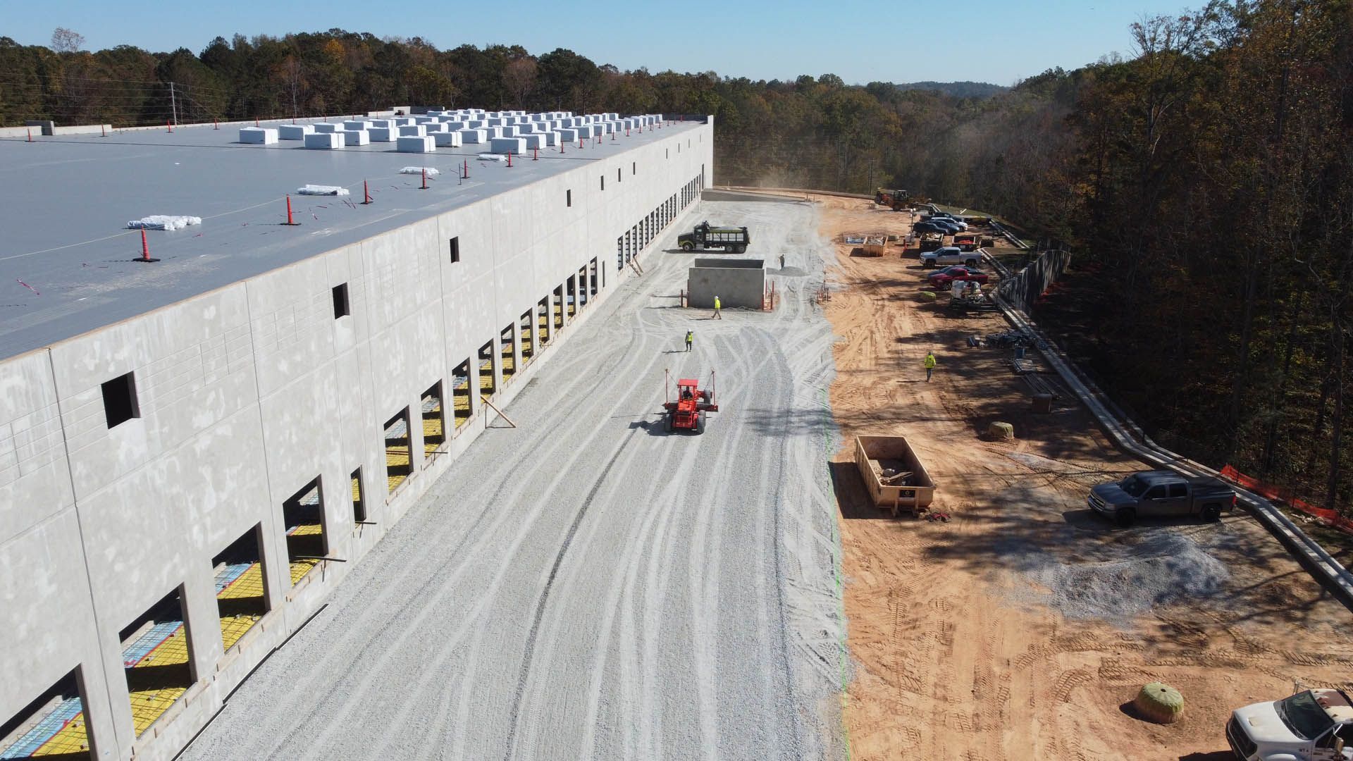 Construction site: large gray building, gravel road, orange lift, trucks, trees, and blue sky.