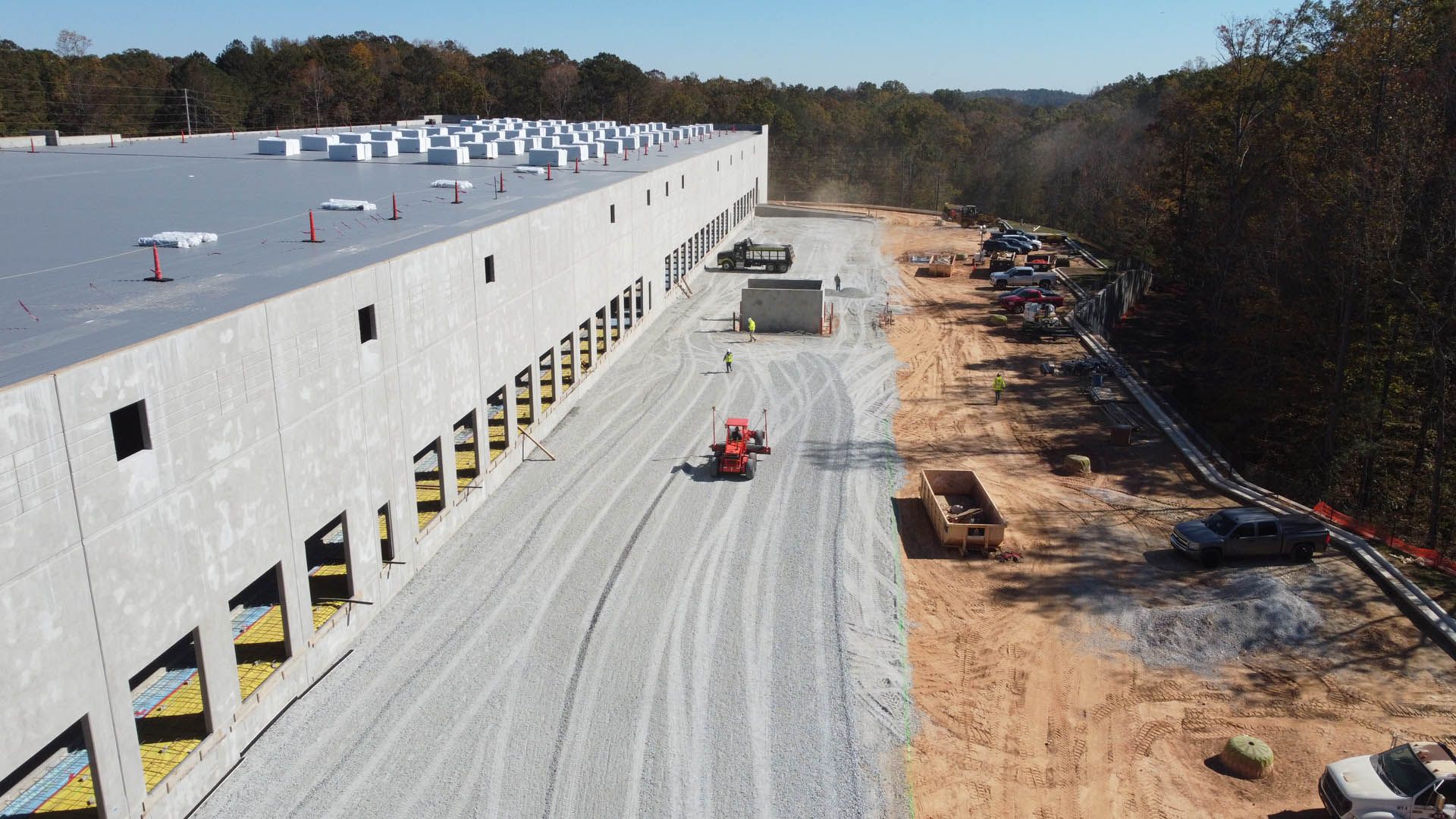 Construction site with a long, light-gray warehouse and gravel road, orange tractor, and parked cars.