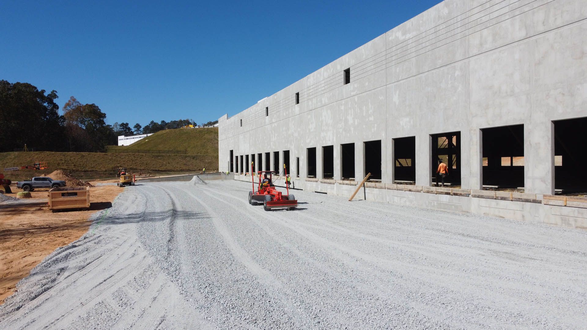 Construction site: building with open loading docks, gravel road, mini roller compactor, blue sky.