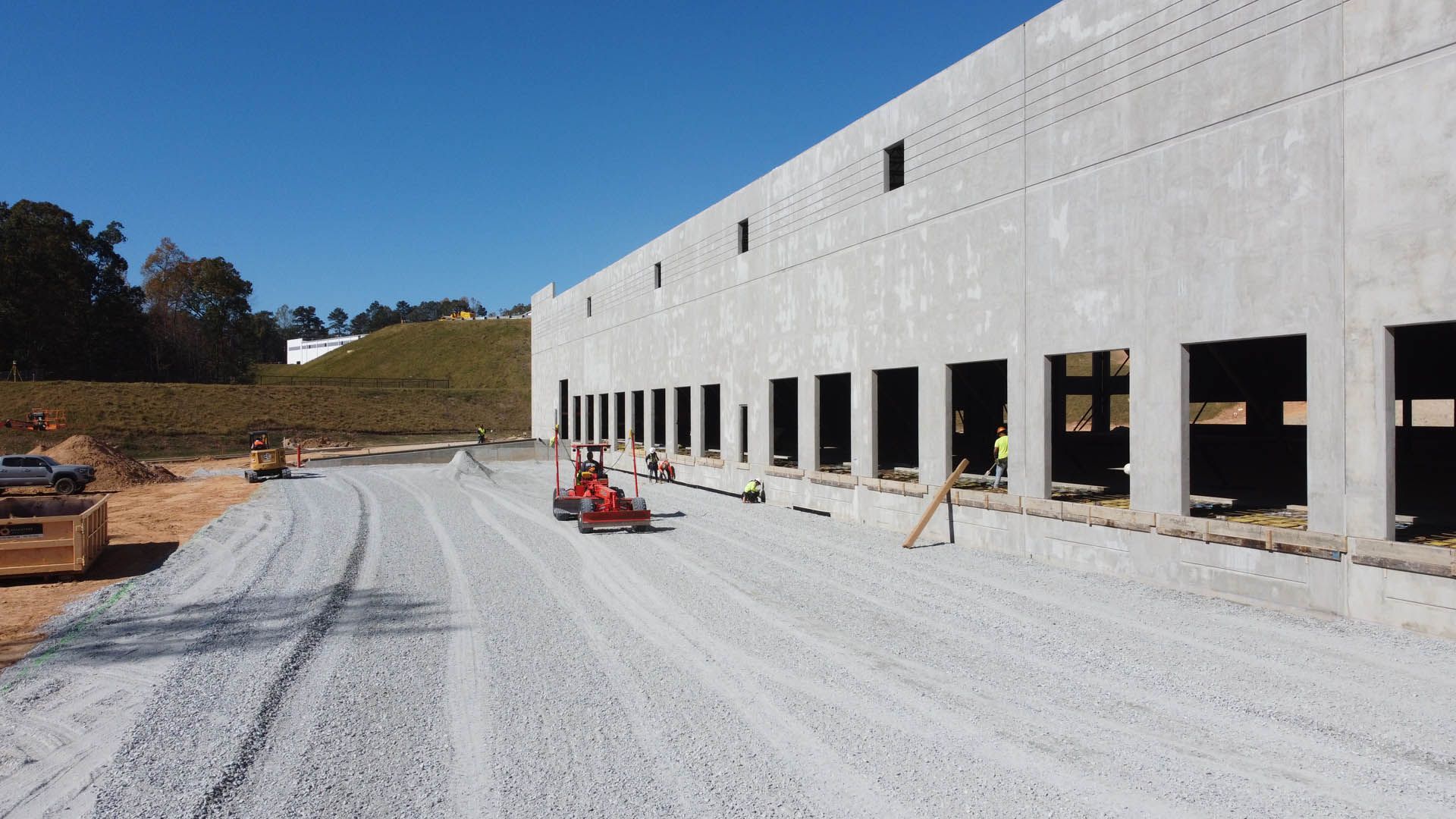 Construction site with a long concrete building and gravel road, bright blue sky.