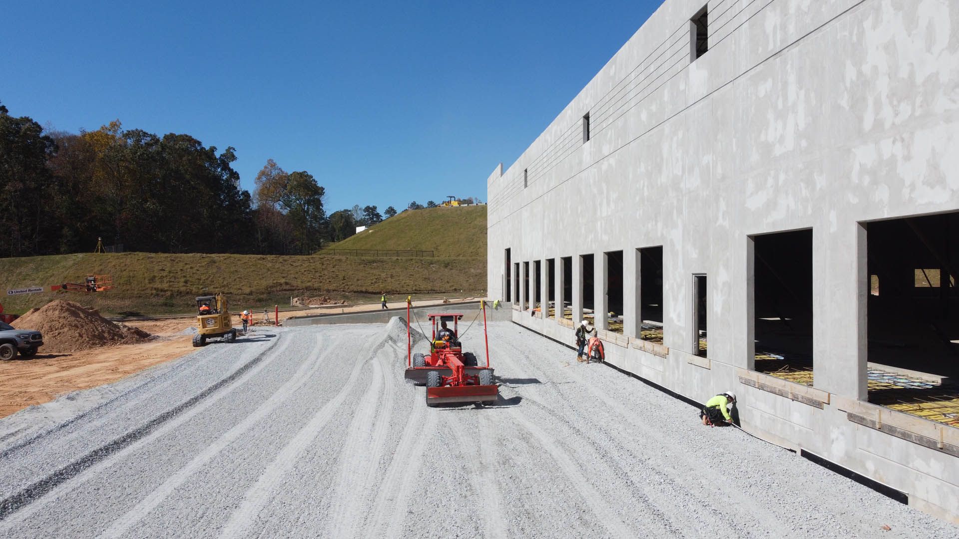 Construction site with paving machinery laying gravel near a concrete building on a sunny day.