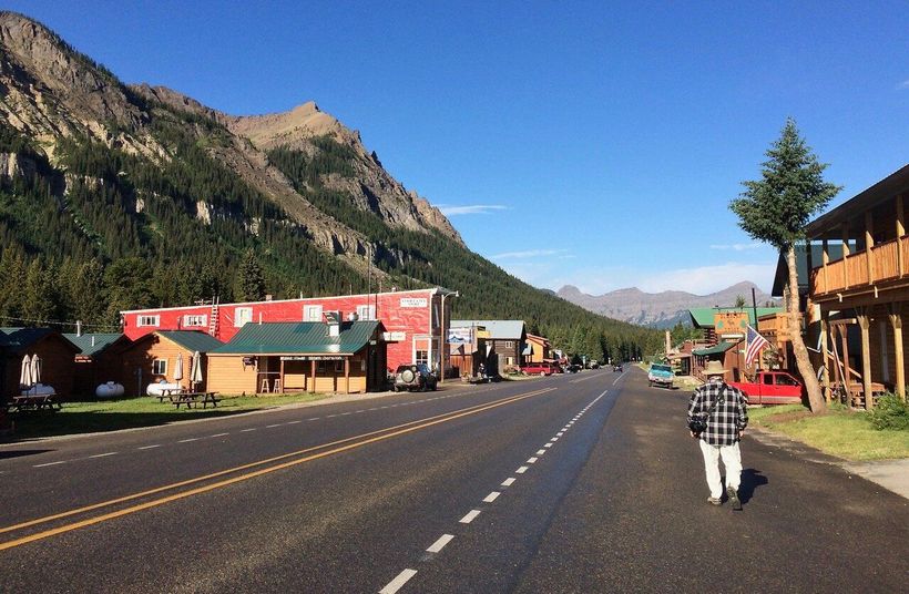 Mountain town street with red storefronts, rocky cliffs, and a person walking on the sidewalk under a clear blue sky.