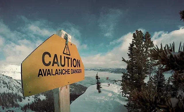 Yellow caution sign warns of avalanche danger on a snowy mountain trail with trees and cloudy sky