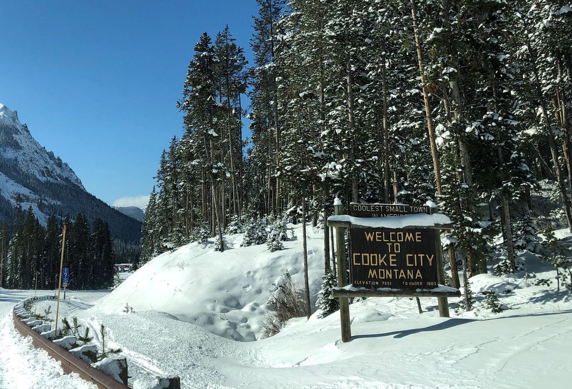 Snowy mountain road with a welcome sign for Eagle Lake, Montana, beside tall pines.