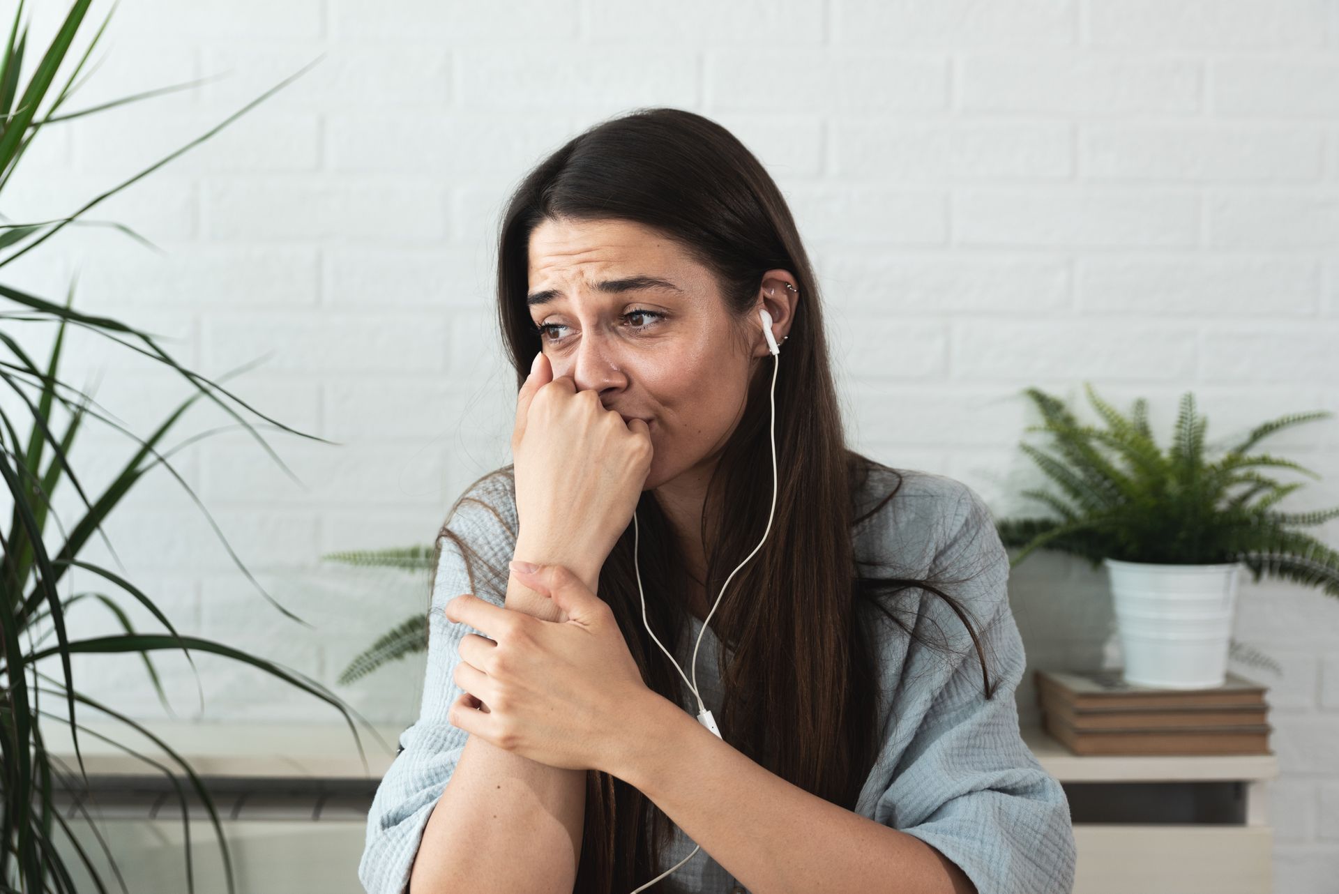 Woman sitting indoors, wiping tears while wearing earbuds, with houseplants and books in the background