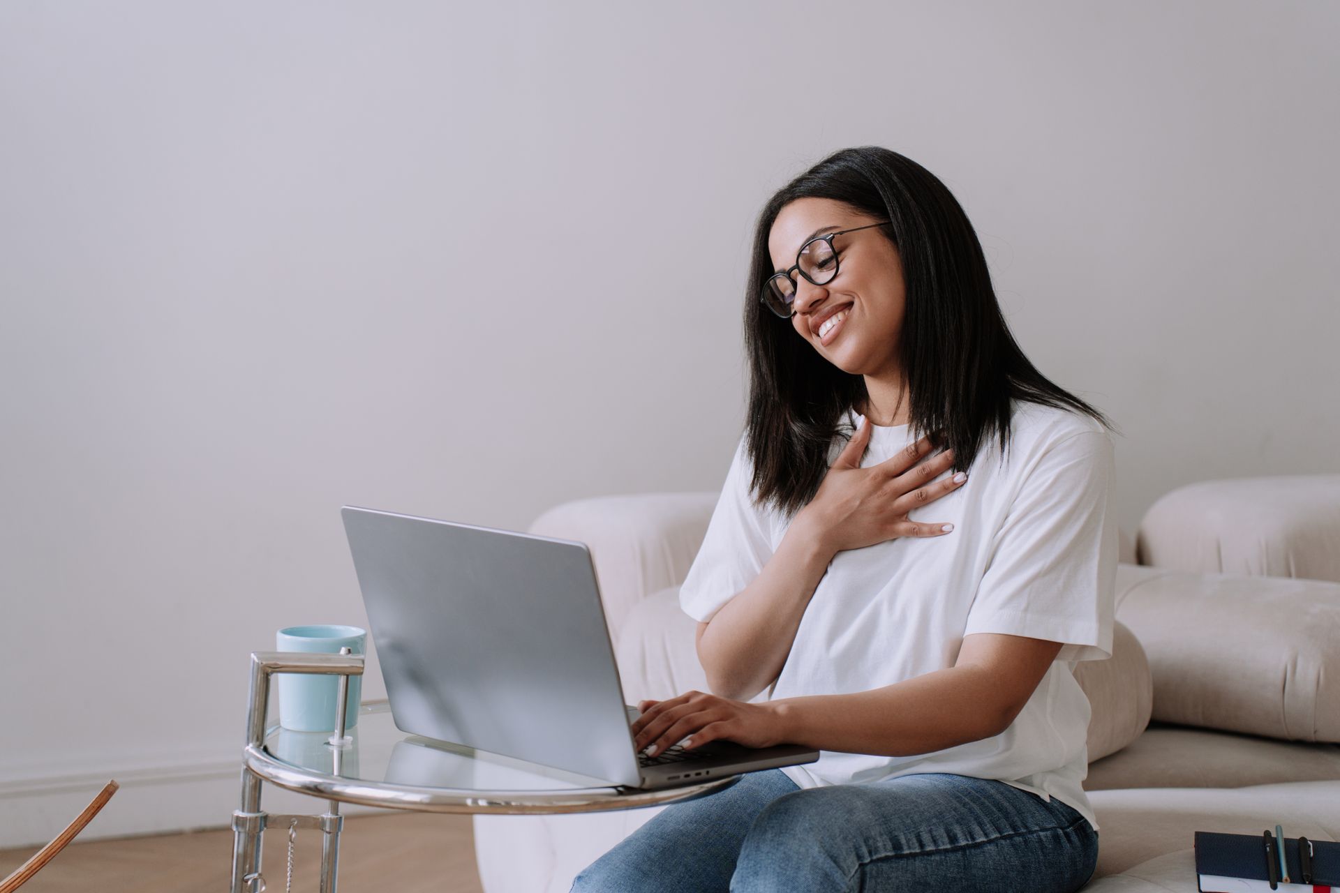 Woman with glasses using a laptop on a sofa, smiling with a hand on her chest in a bright room