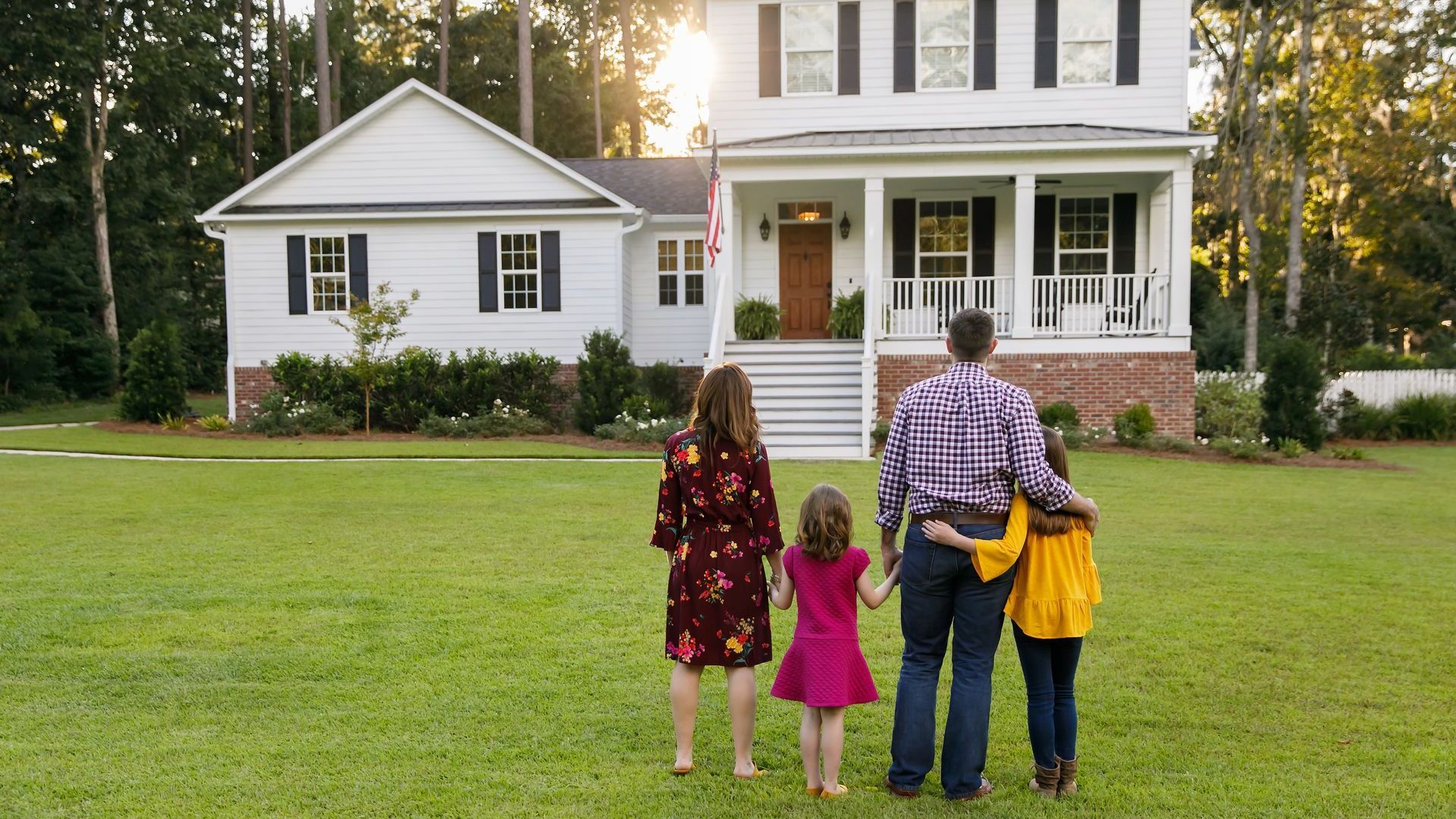 Family of four stands on lawn, looking at two-story white house with shutters and porch.