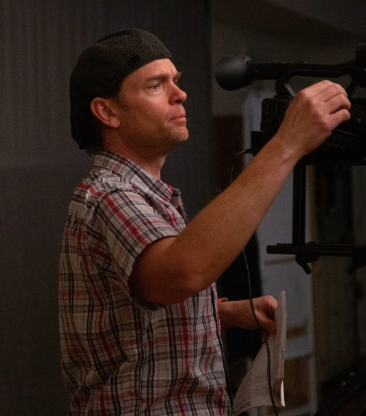Man in a plaid shirt adjusting a TV or camera on a stand indoors